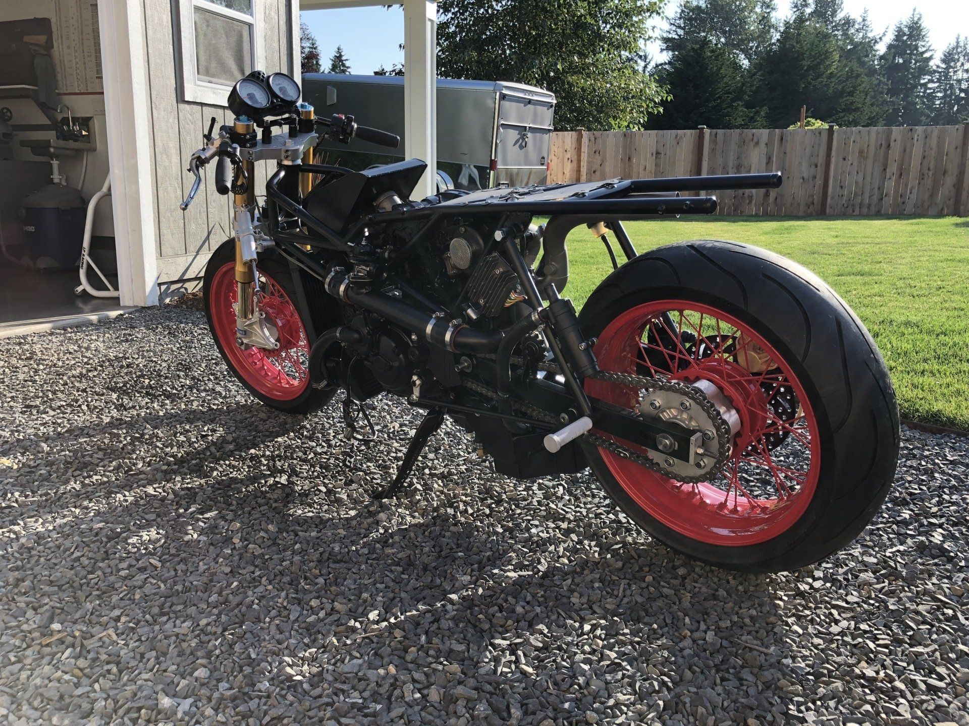 Custom black motorcycle with red wheels parked on gravel beside a house.