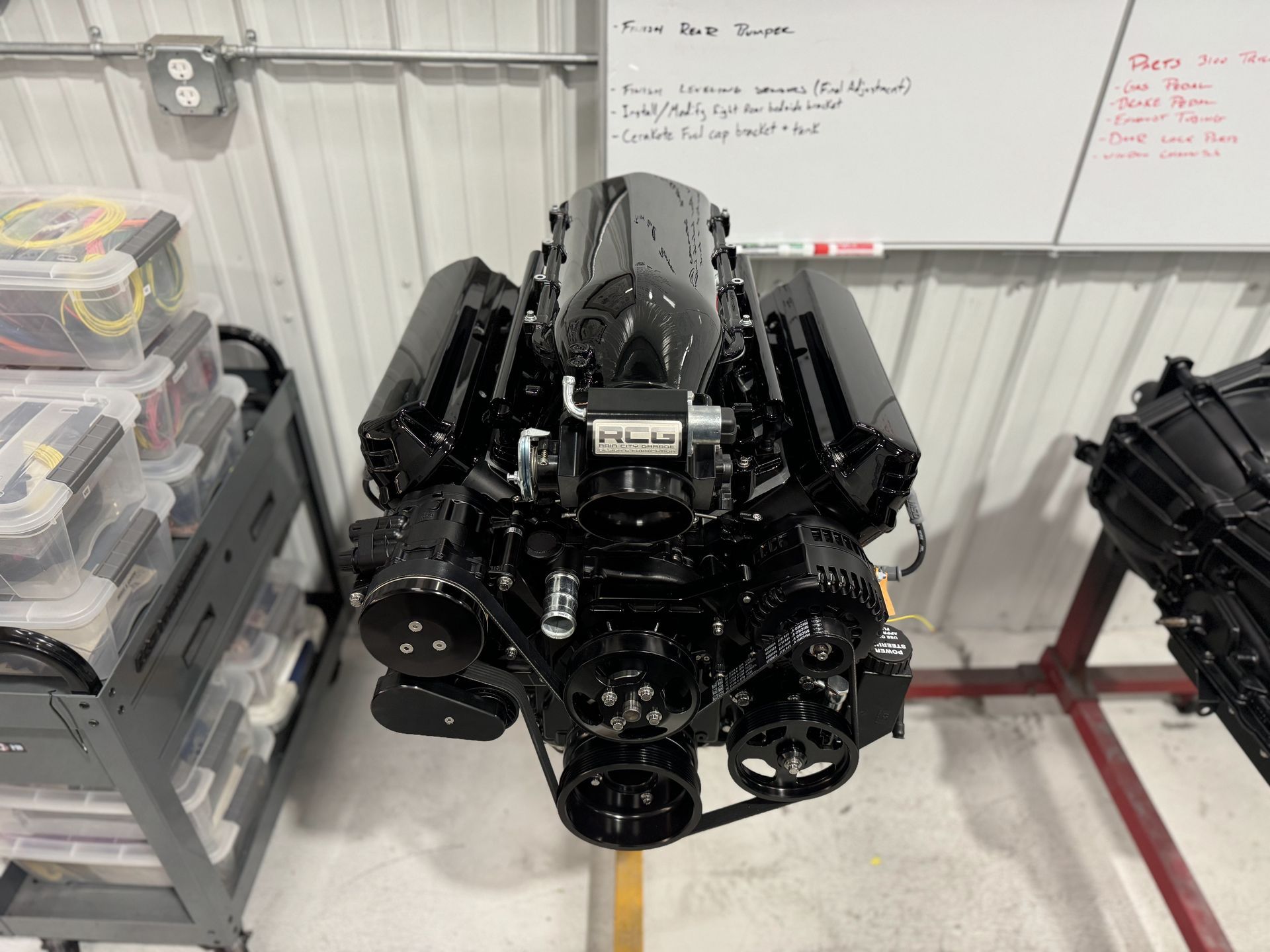 Black engine block on a shop floor between parts bins and a whiteboard in a garage.