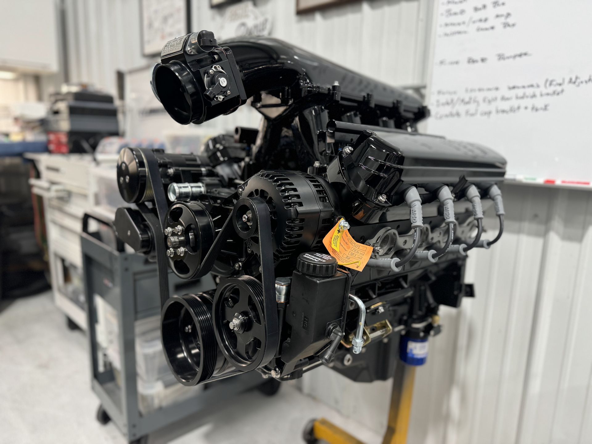 Black car engine on a stand in a workshop, with belts, pulleys, and exposed components visible.