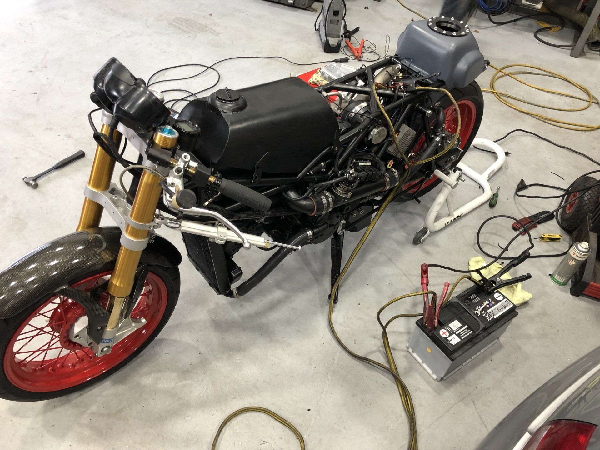 Custom motorcycle with exposed wiring and tools on a garage floor, front red wheel visible.