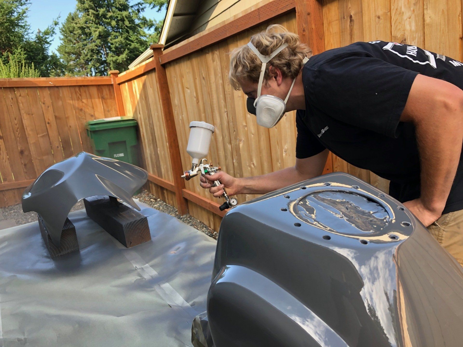 Person spray-painting two gray metal parts on a tarp in a backyard workshop area