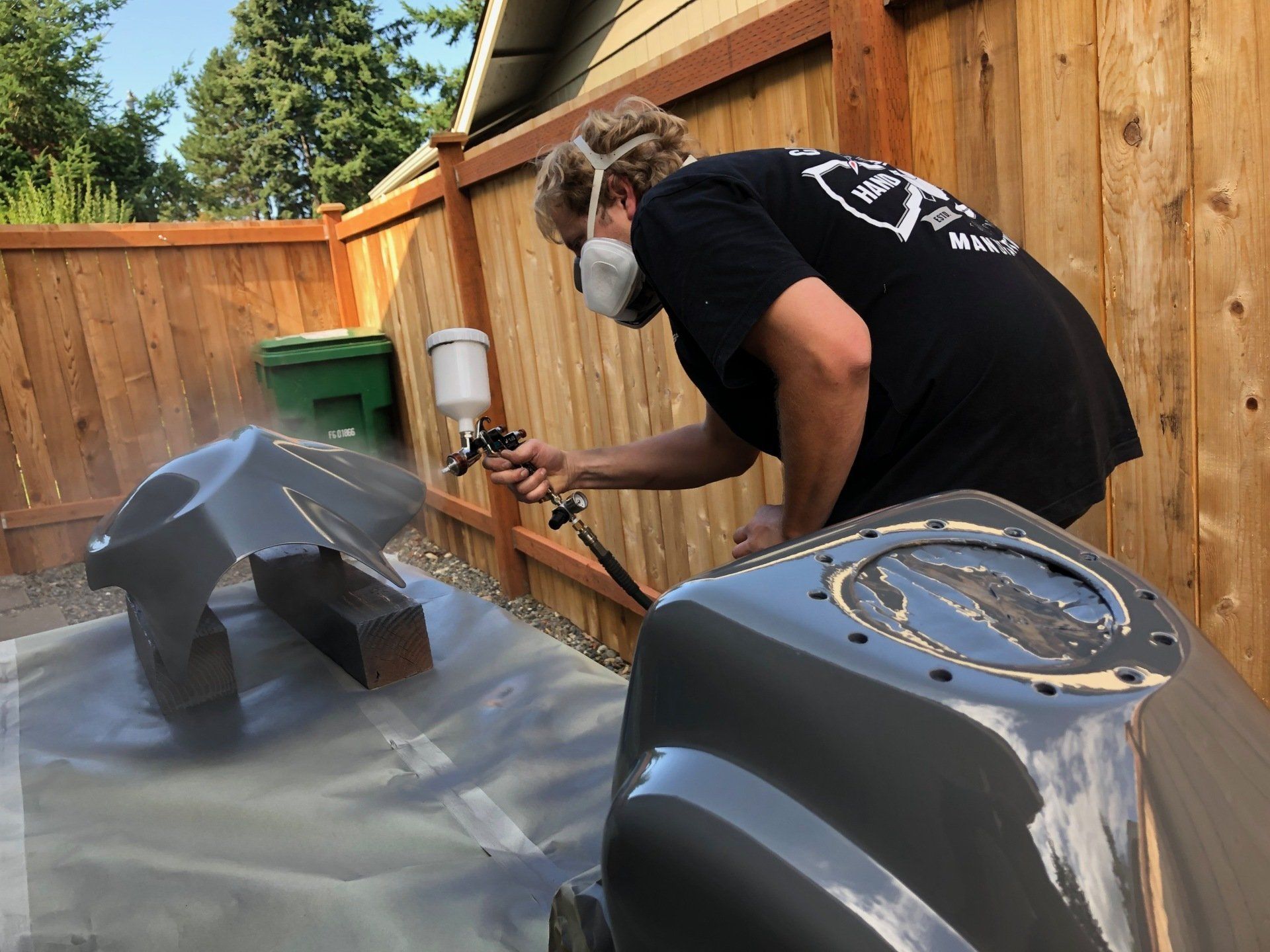 Person spray-painting a gray motorcycle tank in a fenced backyard with a mask on