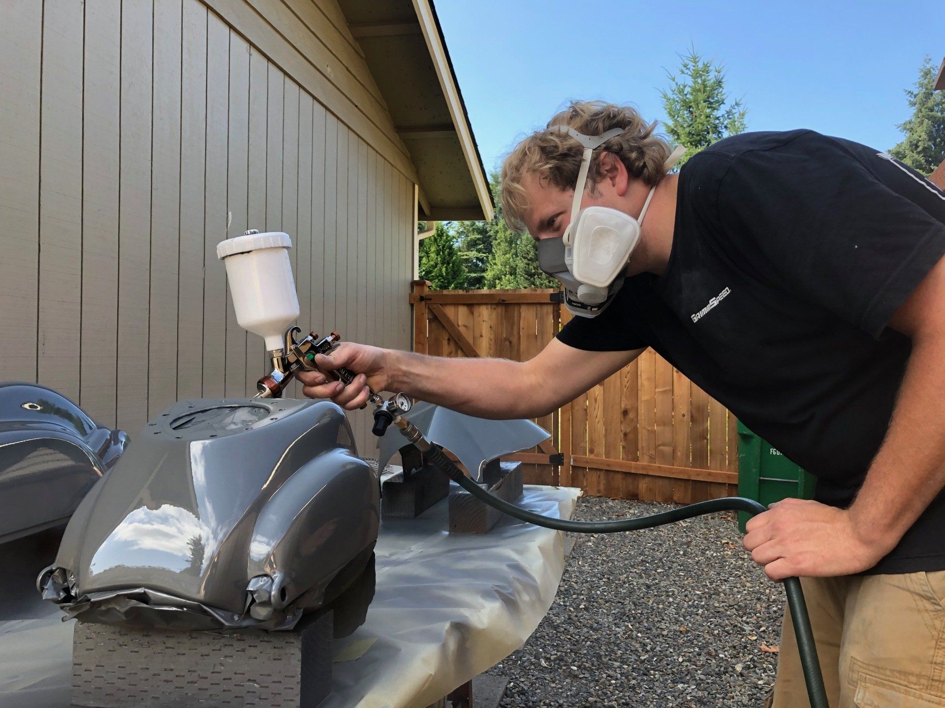 Person in a mask spray-paints a gray motorcycle tank beside a house with a handheld sprayer