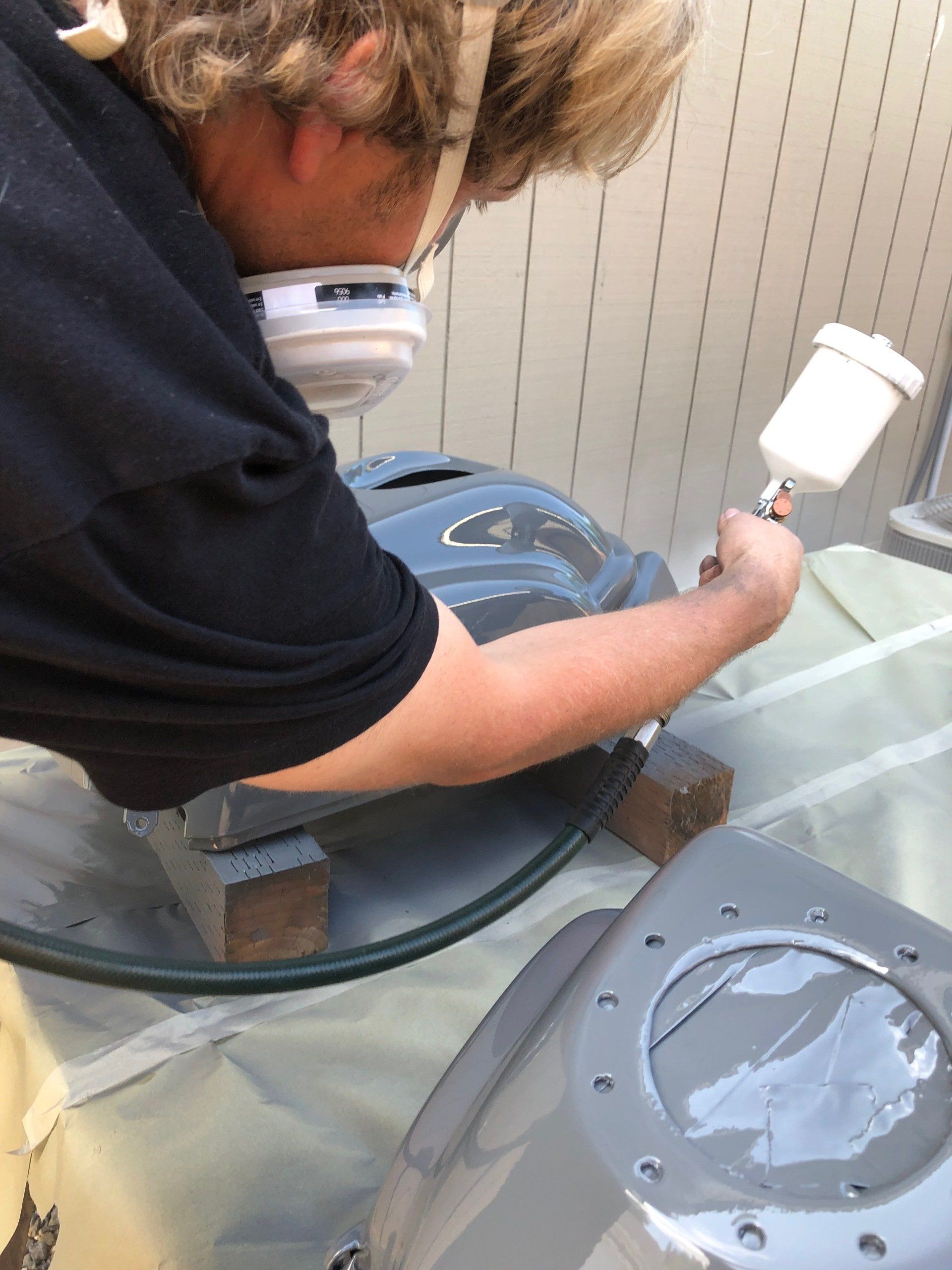 Person wearing a respirator spray-paints a gray object outdoors.