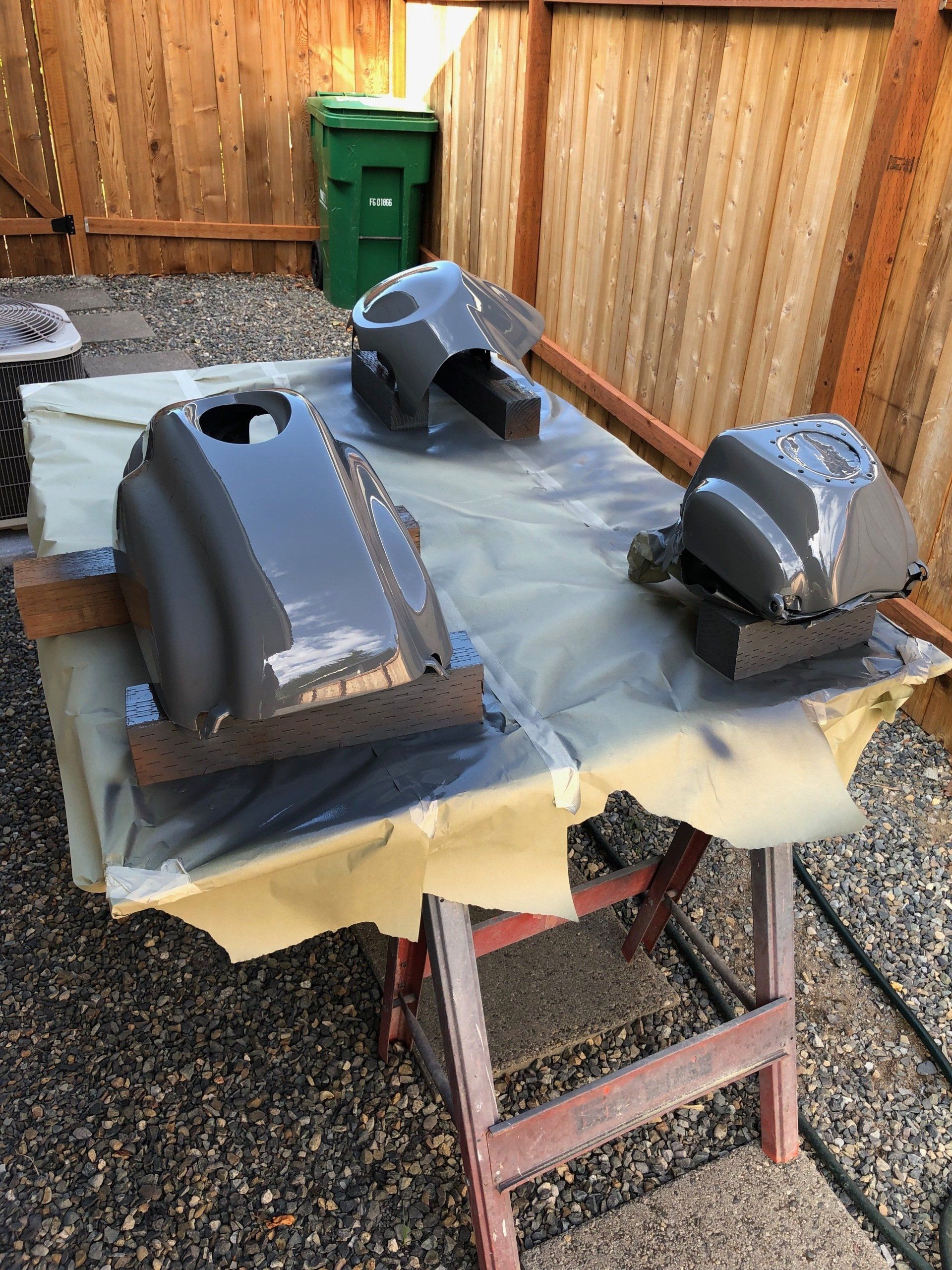 Three gray car body parts drying on a tarp-covered table outdoors near a wooden fence.