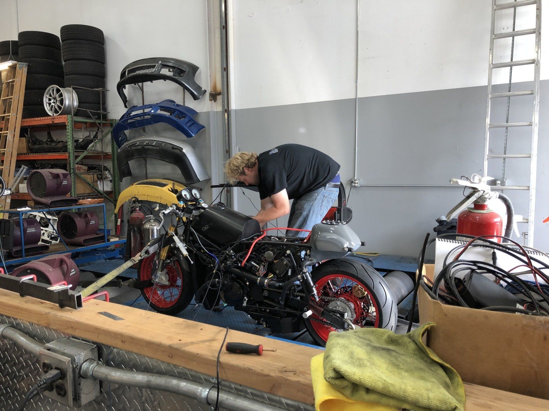 Person bending over a black motorcycle in a workshop with tools, shelves, and a ladder nearby