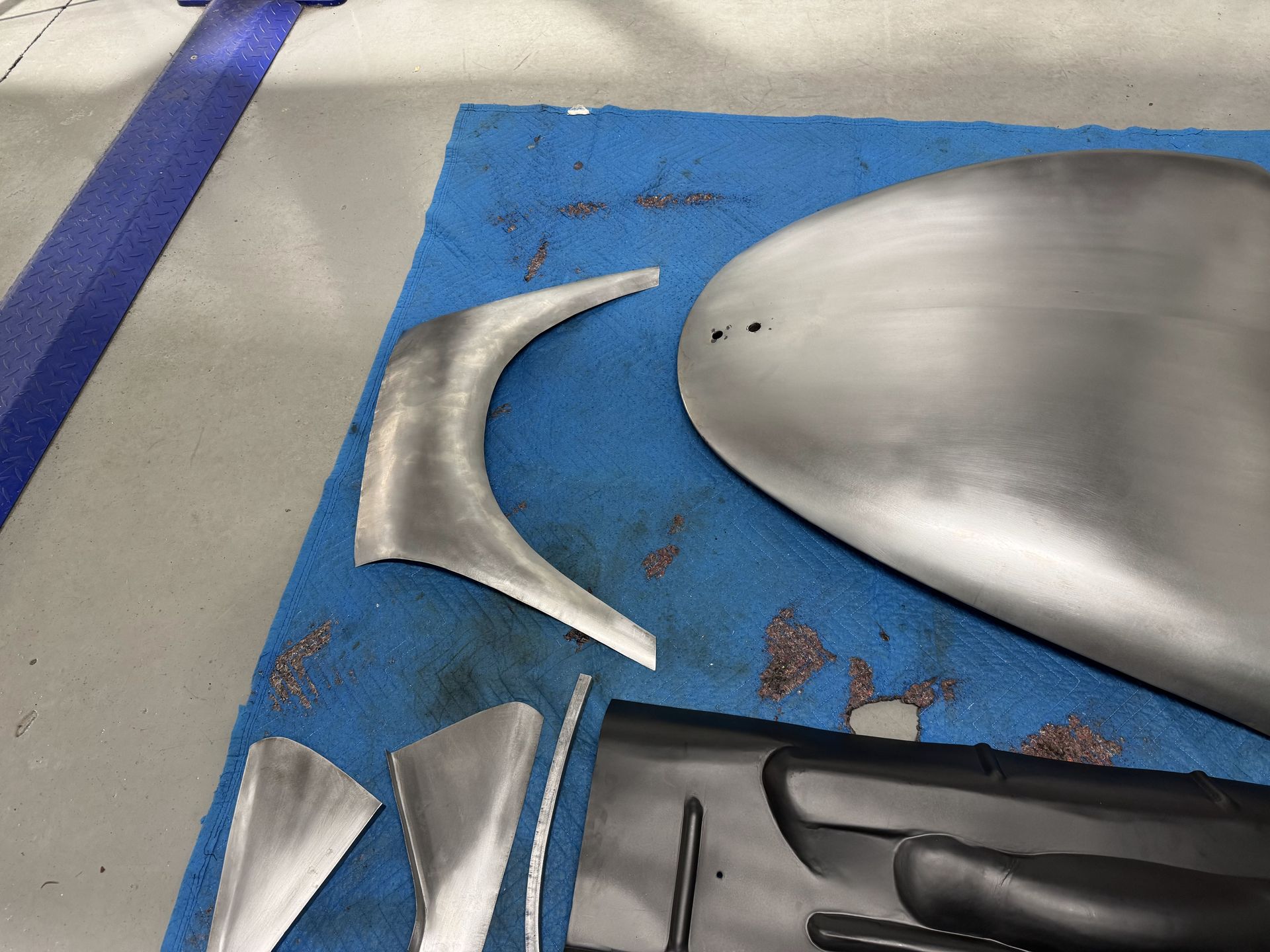 Metal automotive parts arranged on a blue mat, including a curved silver panel and small trim pieces.