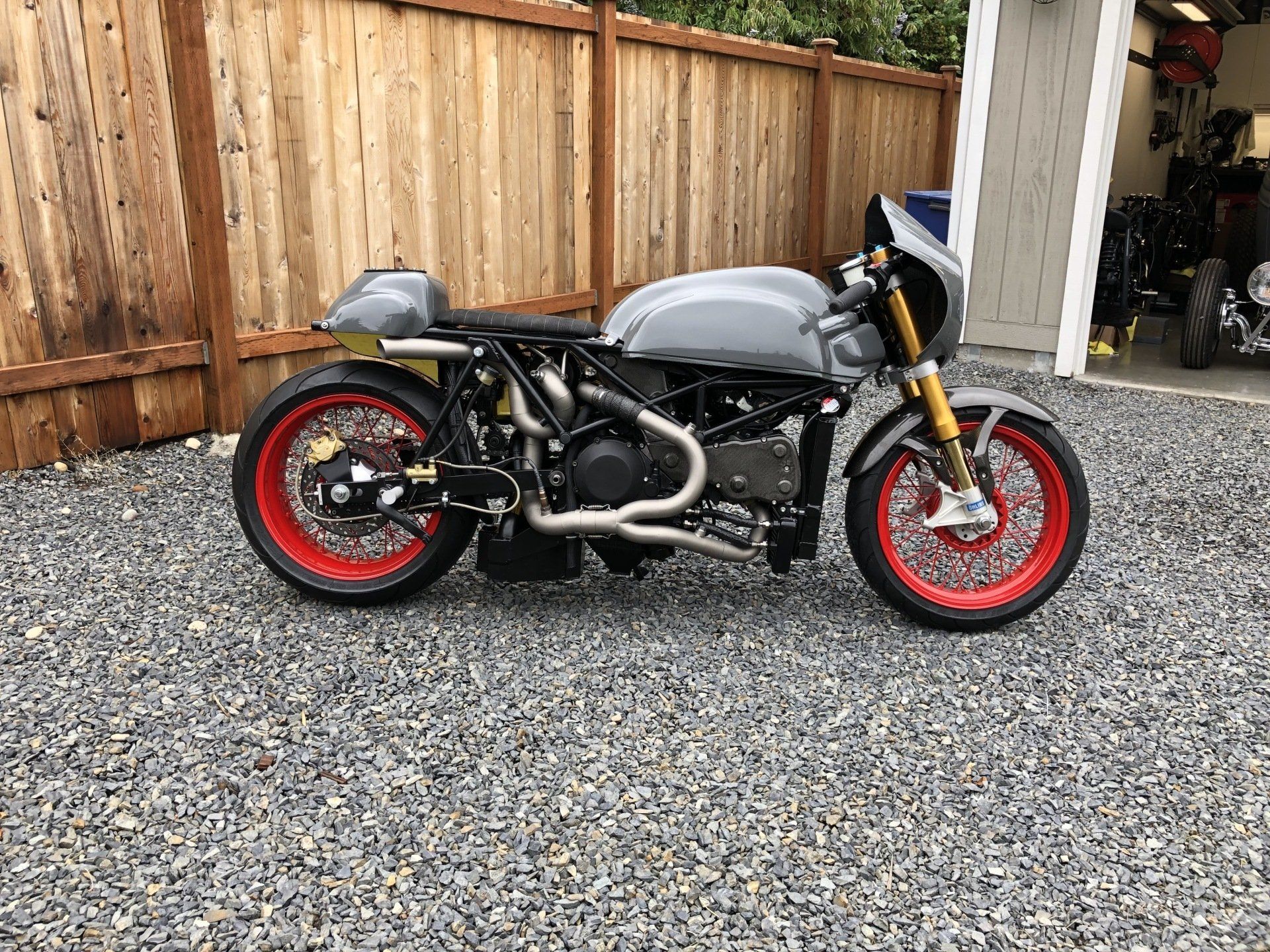 Custom gray motorcycle with red rims parked on gravel beside a wooden fence.