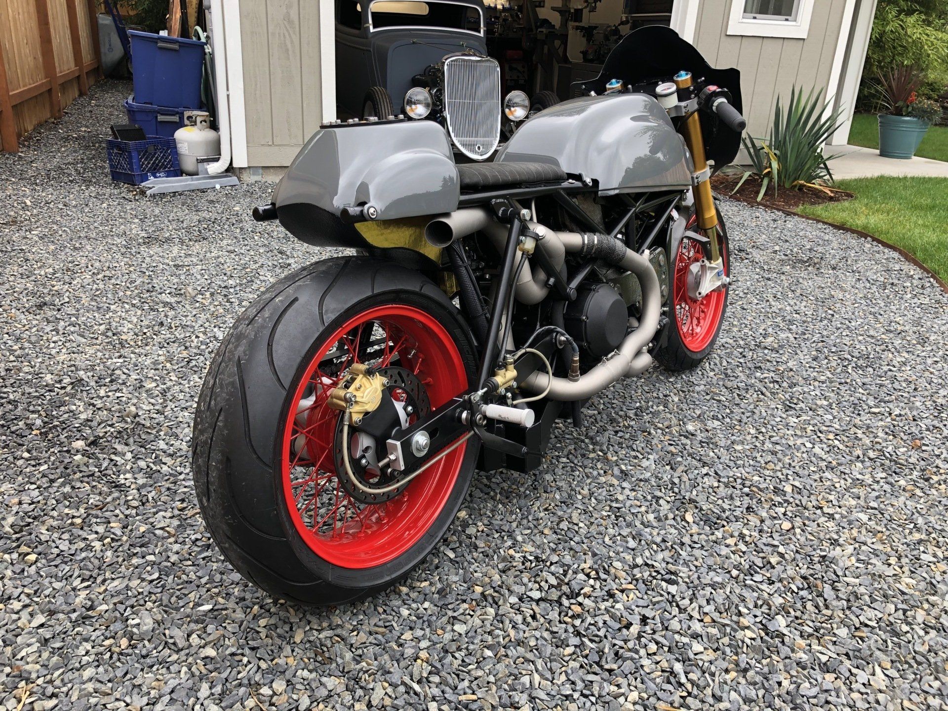 Custom gray motorcycle with red wheels parked on gravel driveway outside a house