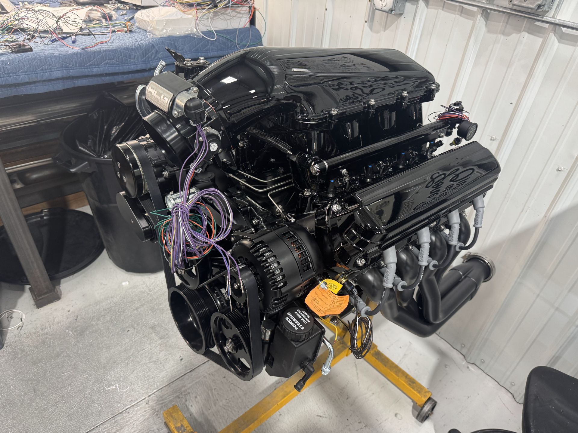 Black car engine on a yellow stand in a garage, with hoses and wiring attached