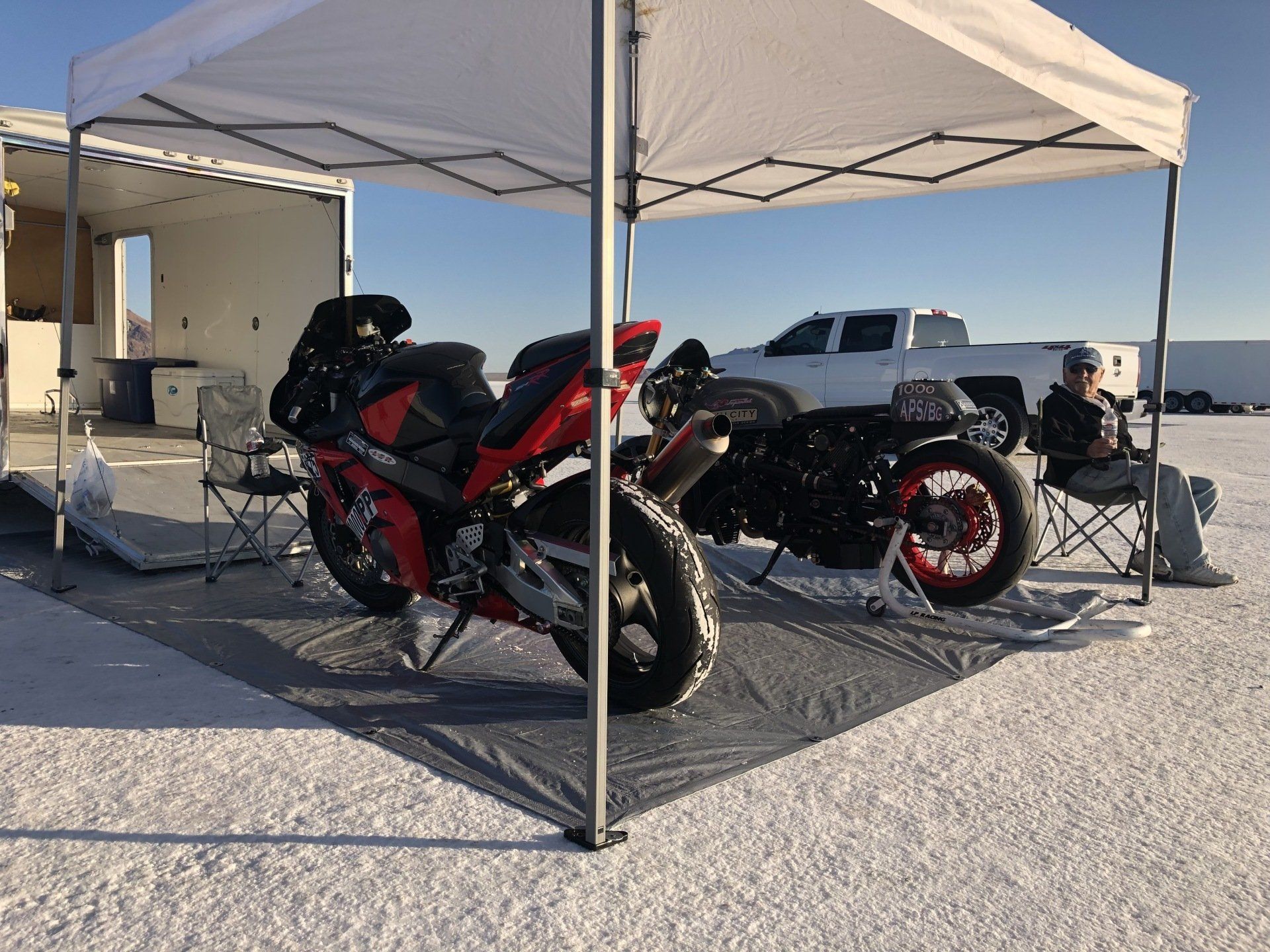 Two red sport motorcycles parked under a white canopy at a gravel campsite, with a pickup truck behind them.