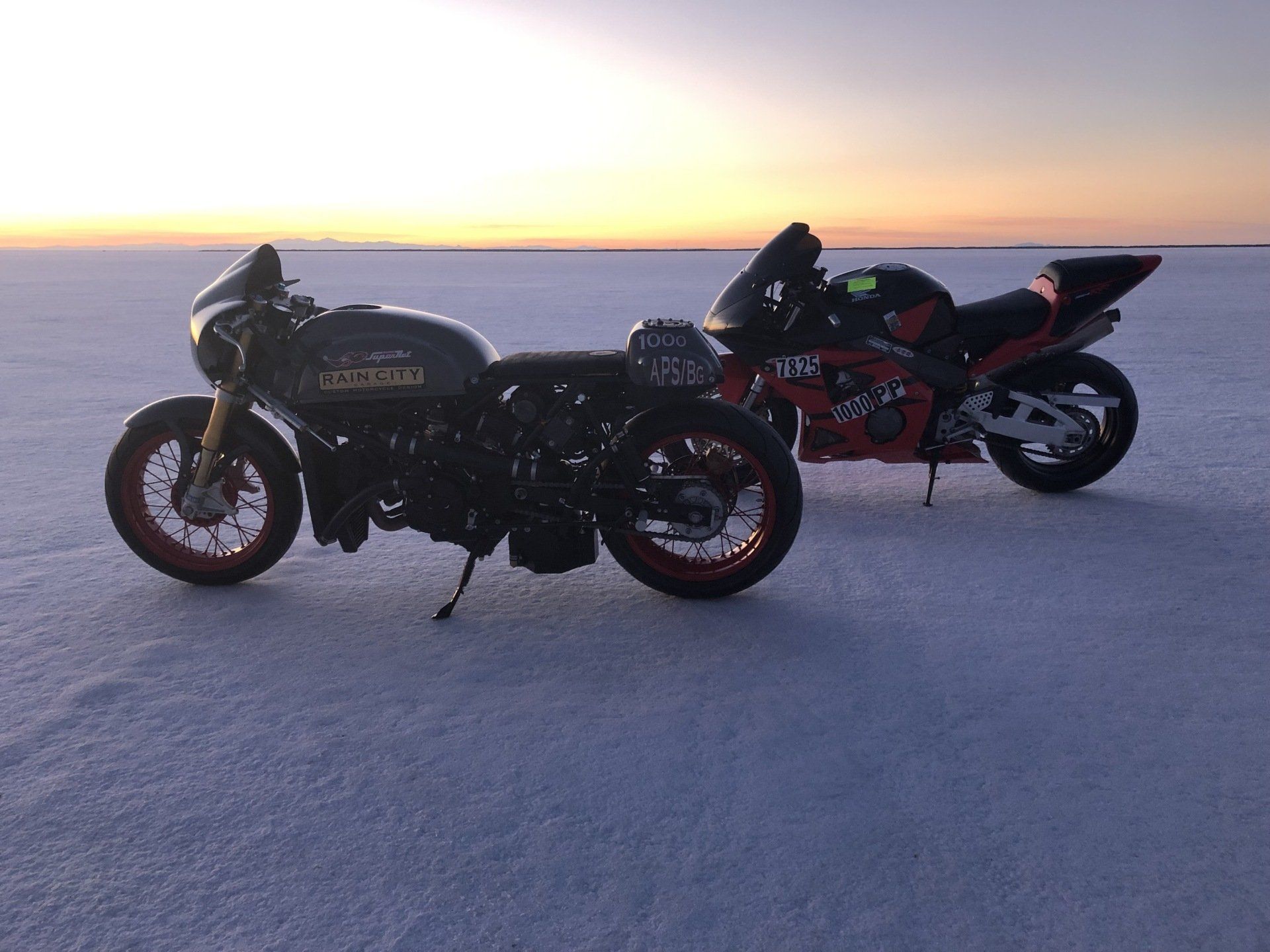 Two motorcycles parked on a snowy plain at sunset, with a glowing horizon in the background.