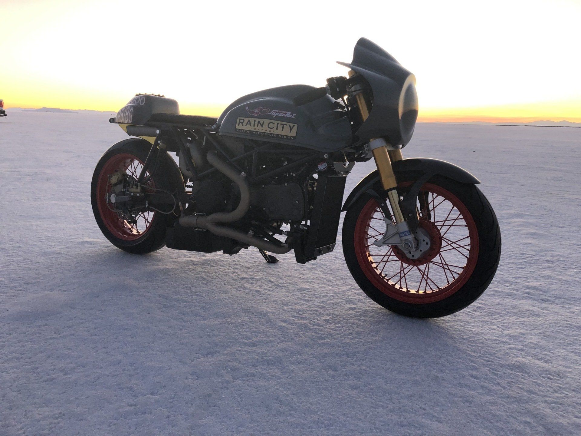 Black custom motorcycle on a salt flat at sunset, with red wheels and a distant horizon.