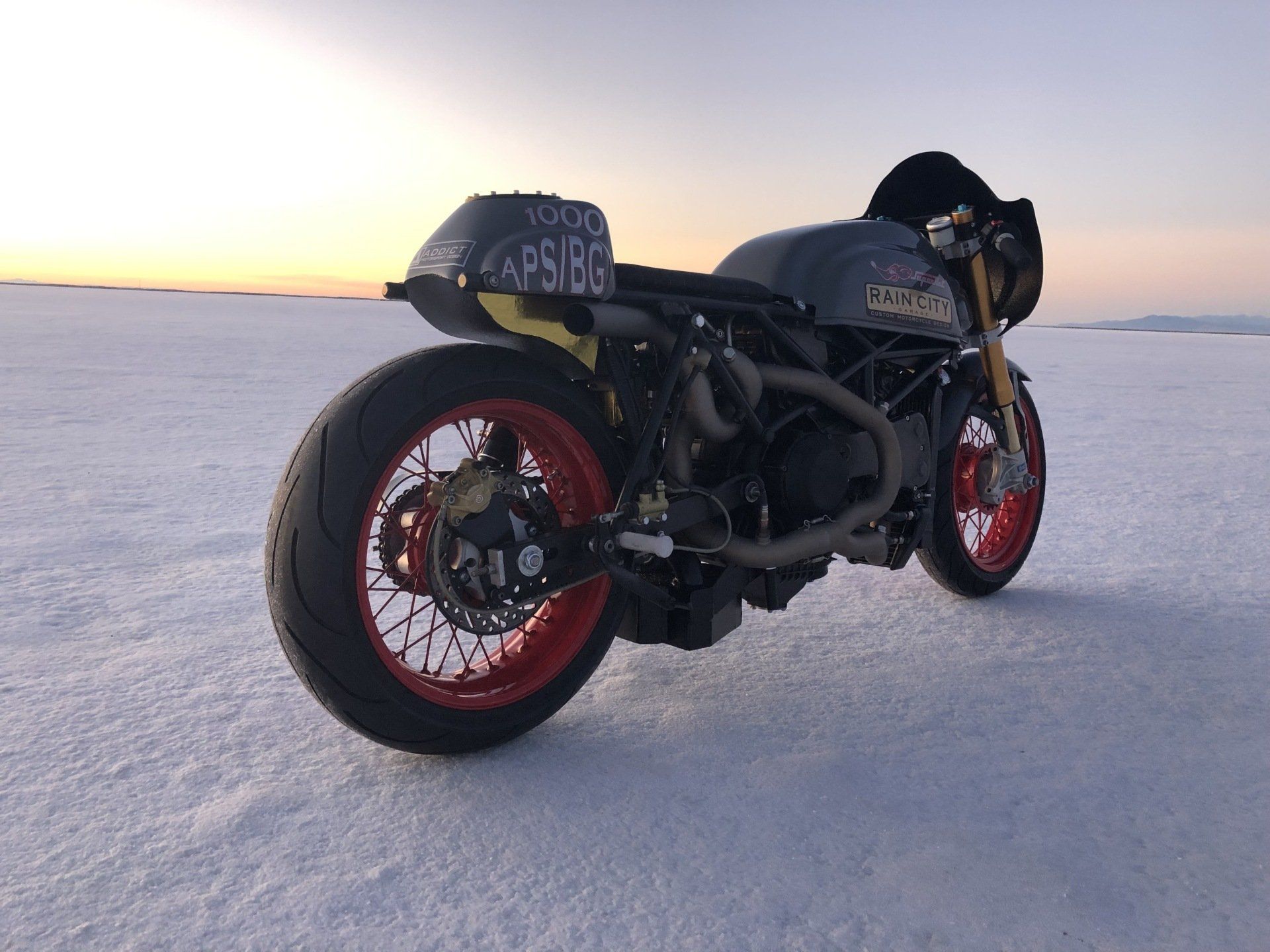 Black custom motorcycle with red rims parked on snowy ground at sunset.