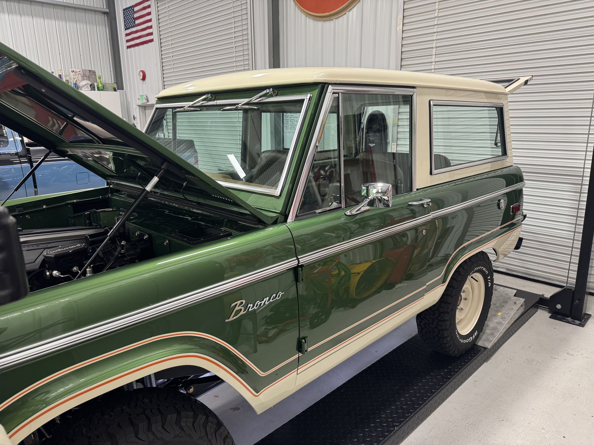 Green vintage Ford Bronco in a garage with its hood open, white roof and tan interior visible.