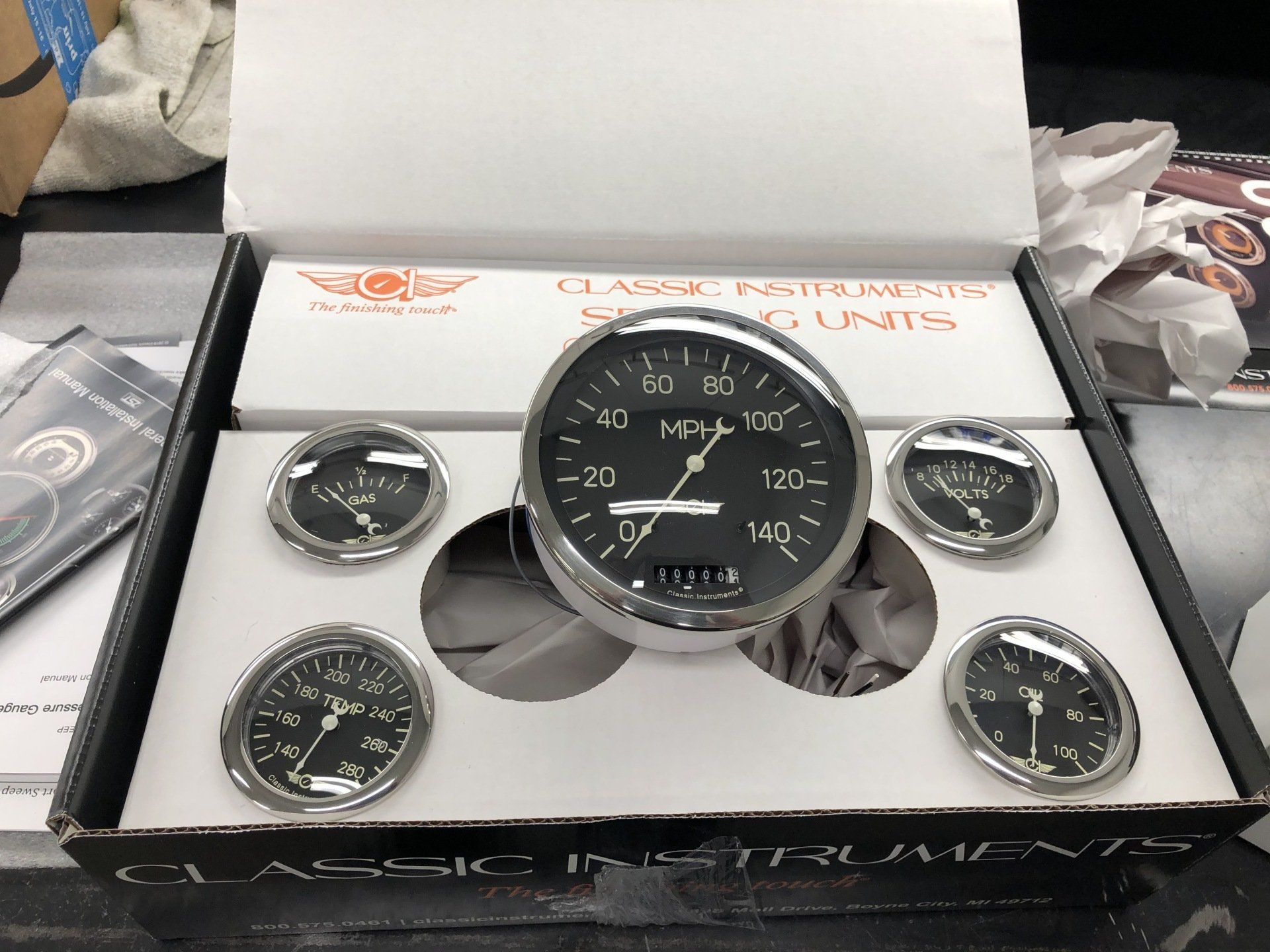 Black CLiQ automotive gauges in a display box, including a large central gauge and four smaller dials.