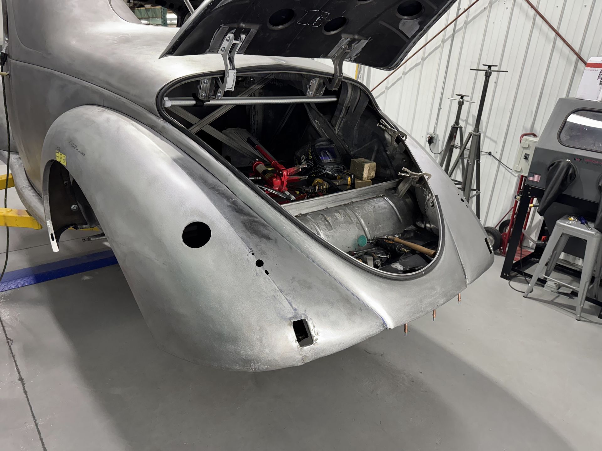 Silver sports car body in a workshop, with the rear hatch open and exposed engine components.