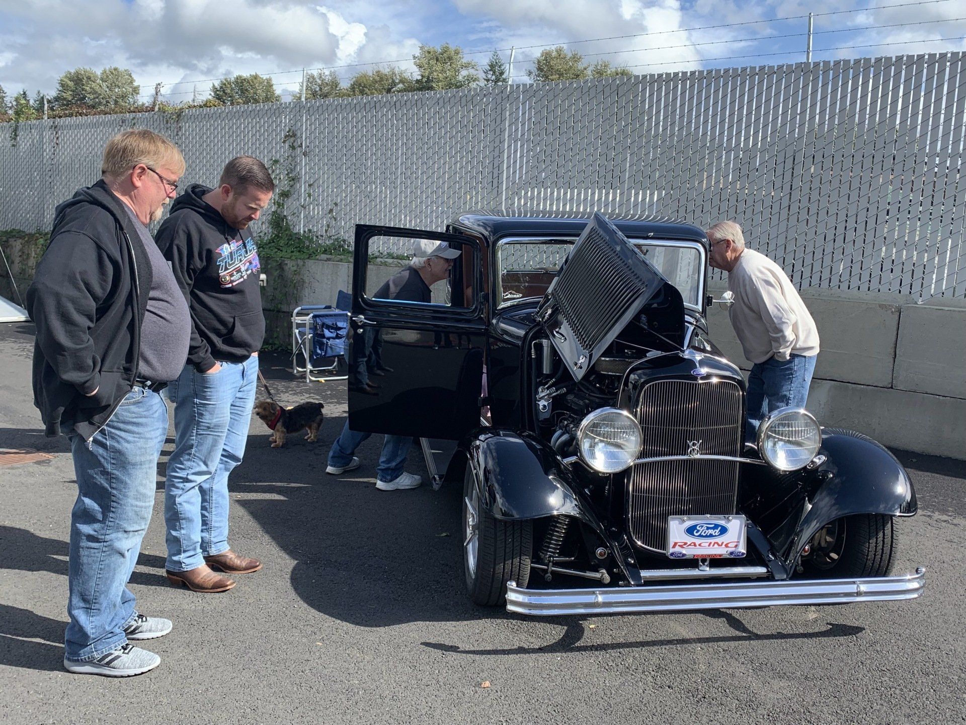 Three men examine a black vintage car with its hood open in a parking lot.