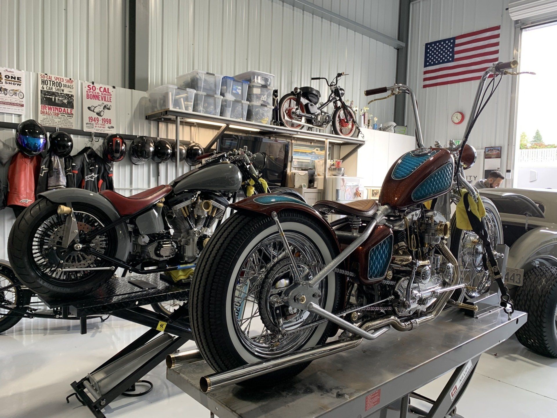 Two custom motorcycles on lifts in a workshop, with an American flag on the wall.