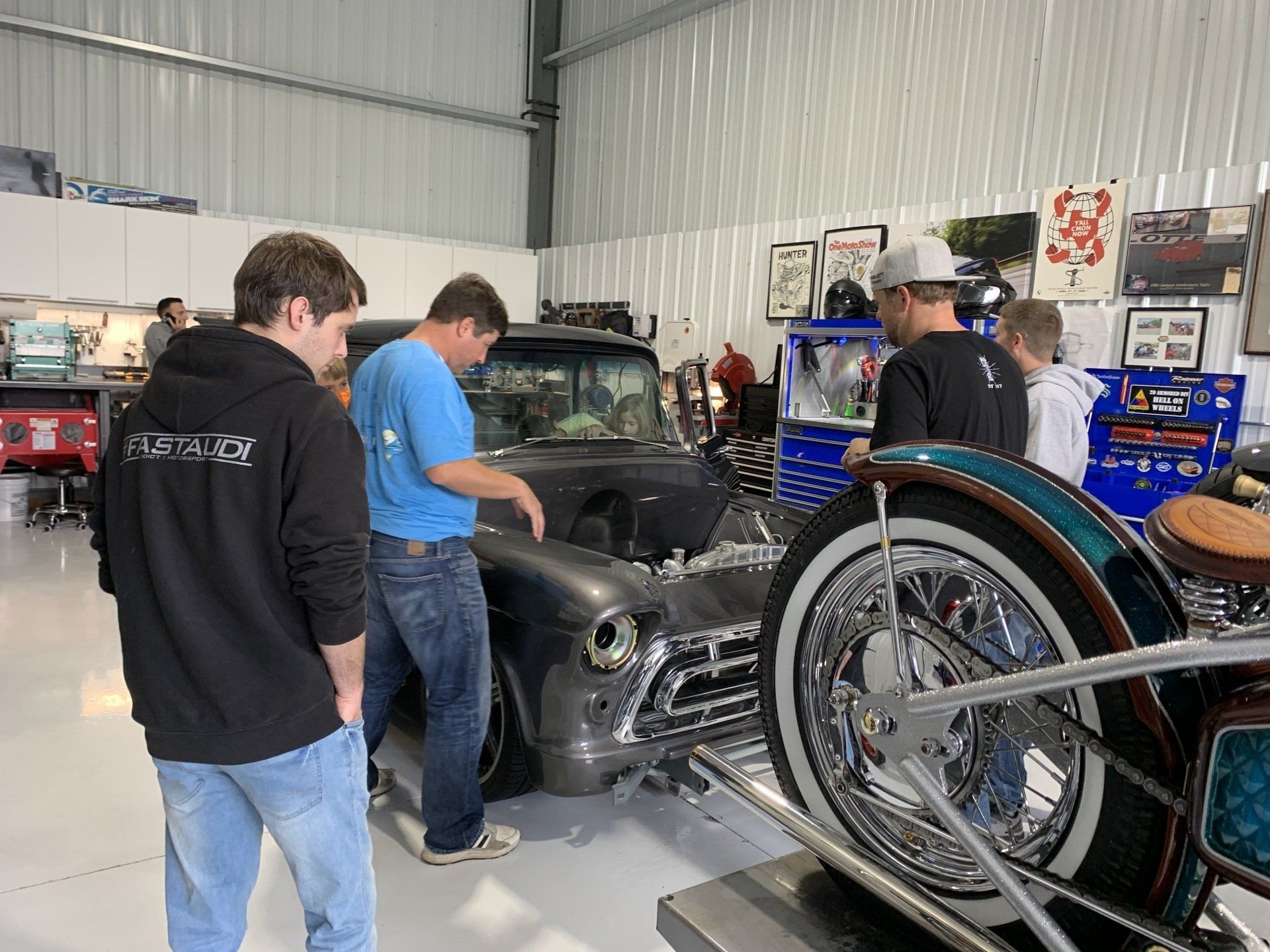 People examining a vintage black car inside a workshop, with a motorcycle wheel in the foreground