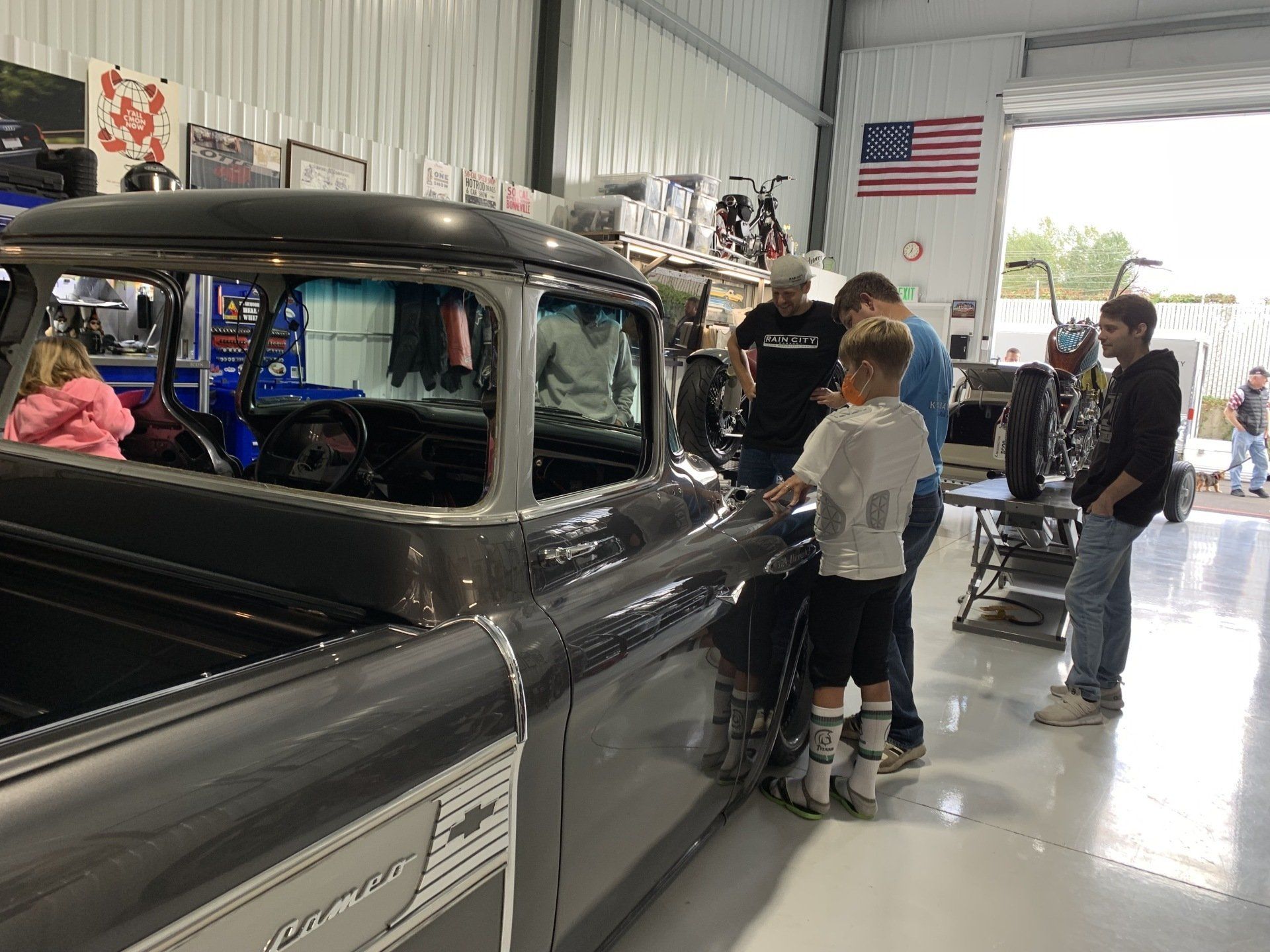Classic black car in a garage with two people looking inside and an American flag on the wall