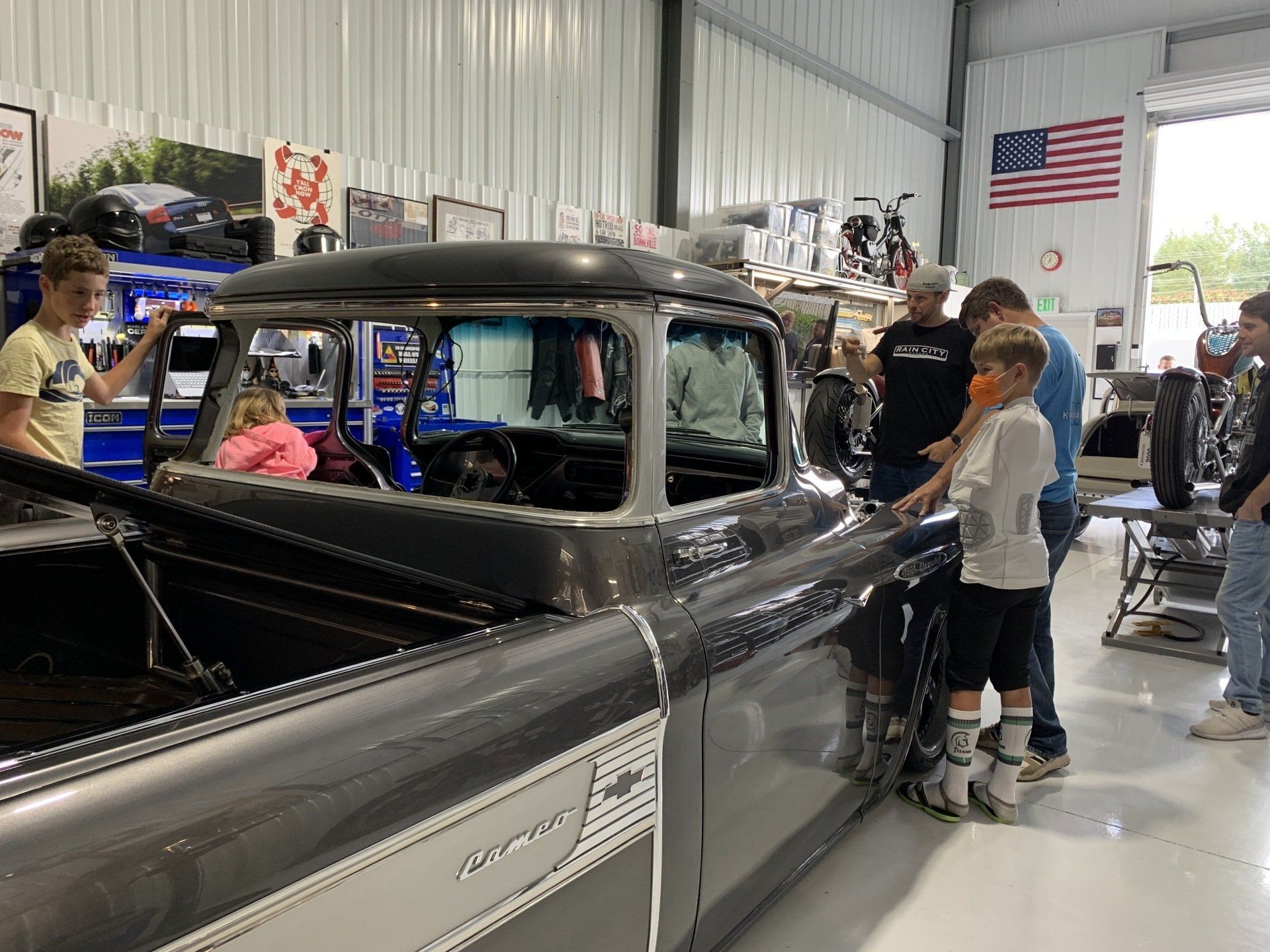 Classic black pickup truck inside a garage with people working on it under an American flag.