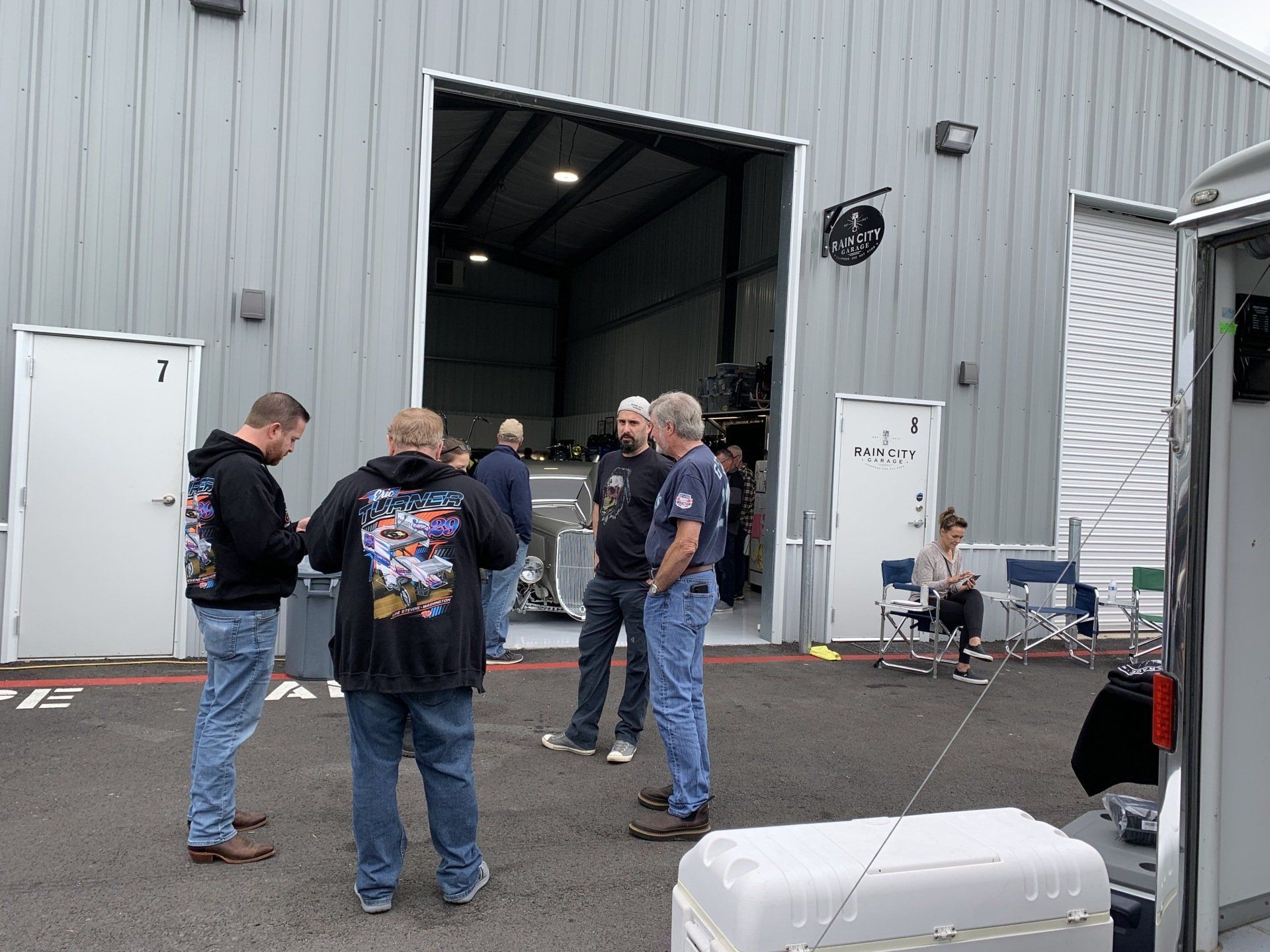 People gathered outside an open warehouse bay, talking beside parked vehicles and equipment.