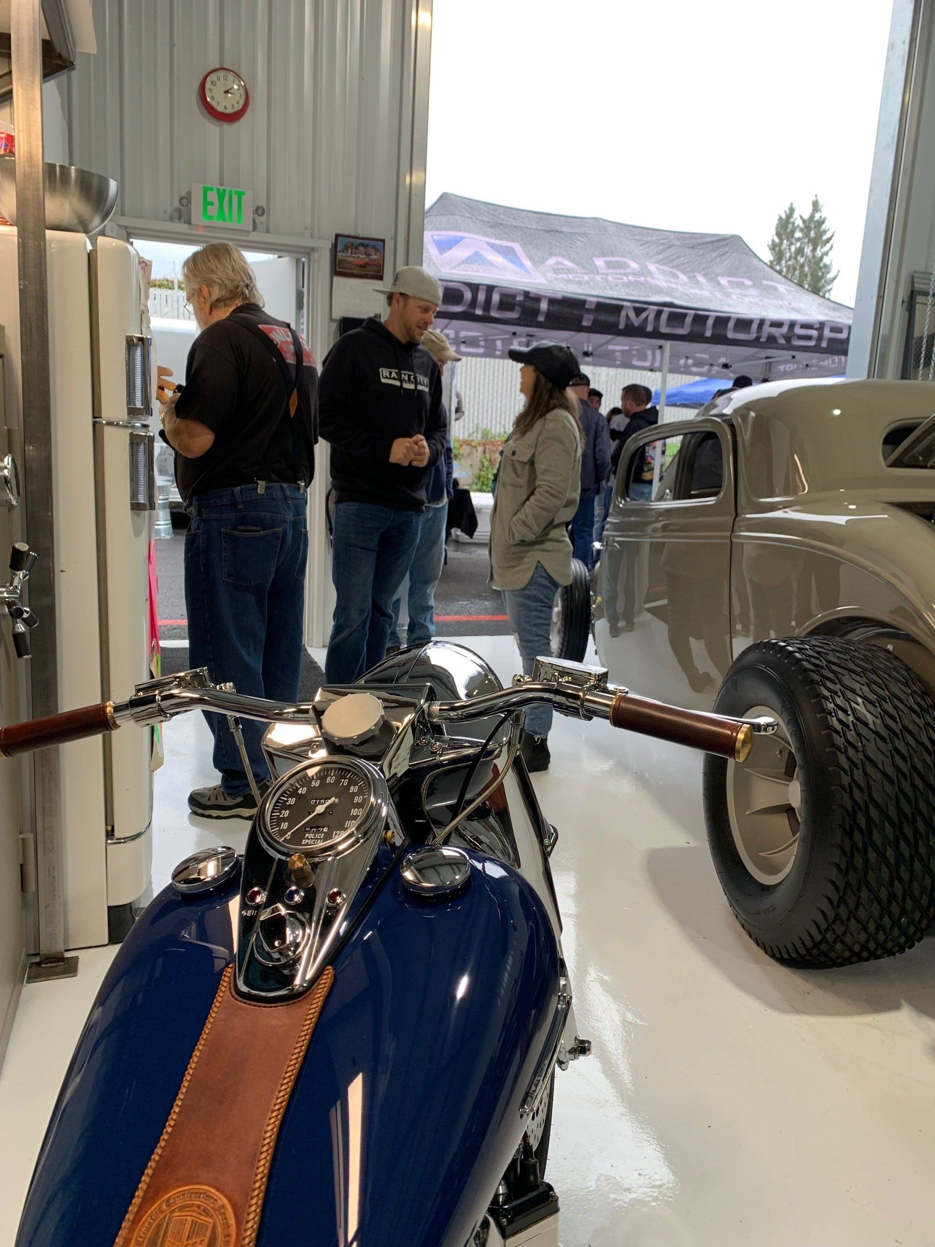 Blue motorcycle in a garage, with people standing near a vintage car and a trailer outside.