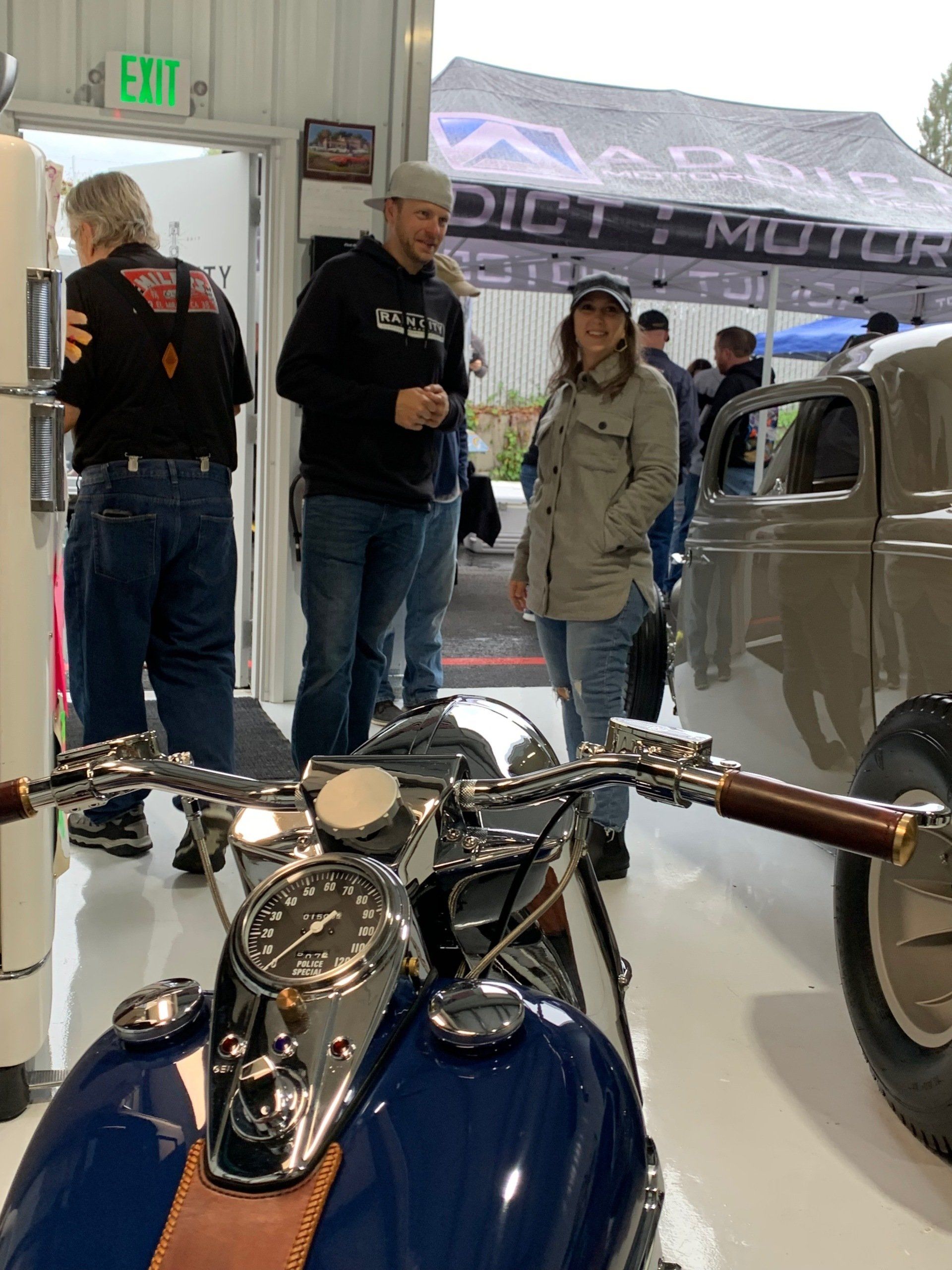 Motorcycle showroom with blue bike in foreground and people browsing near a vintage car and tents outside.