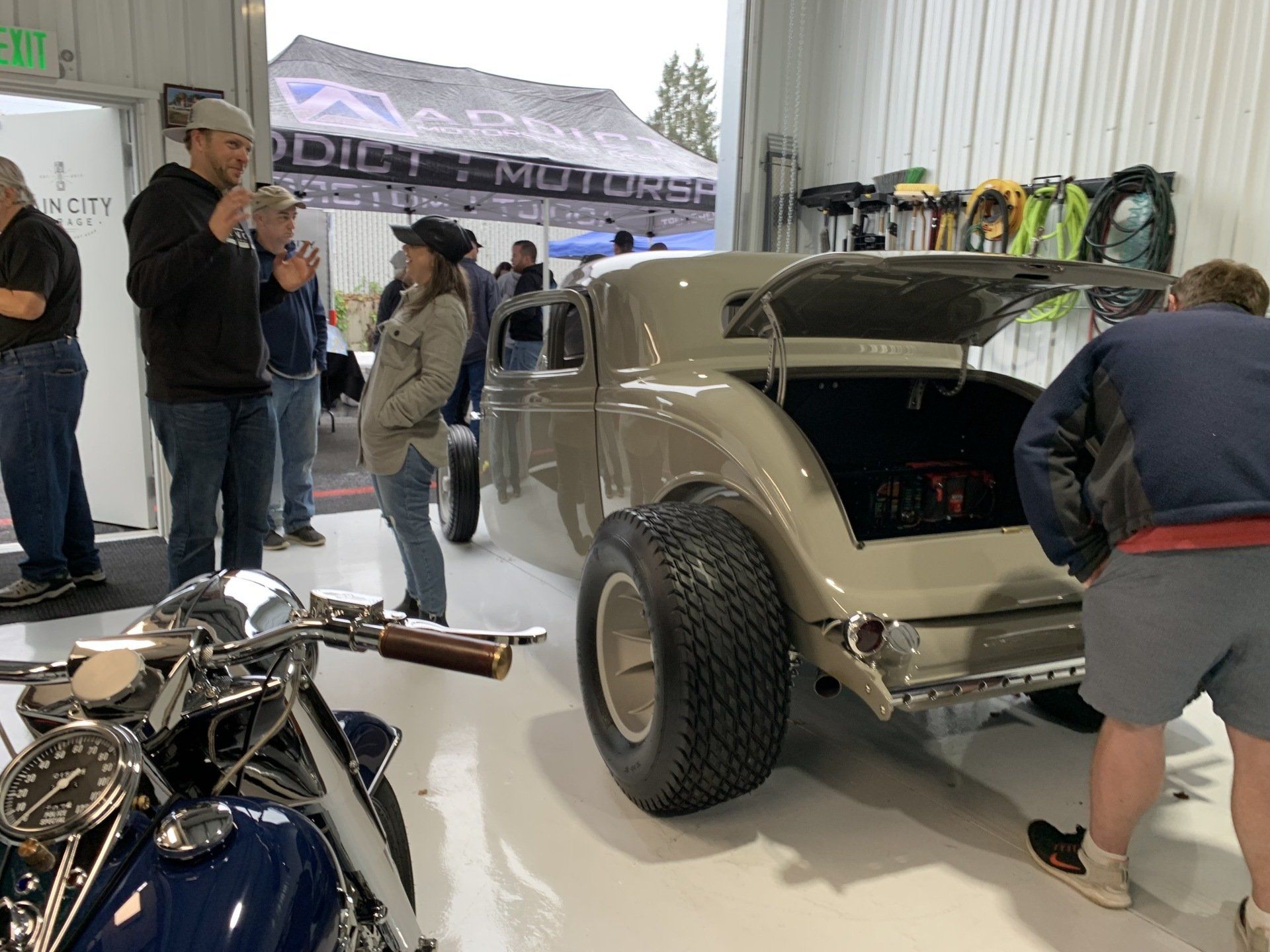 Classic beige hot rod at indoor car show with people gathered around and a motorcycle in the foreground