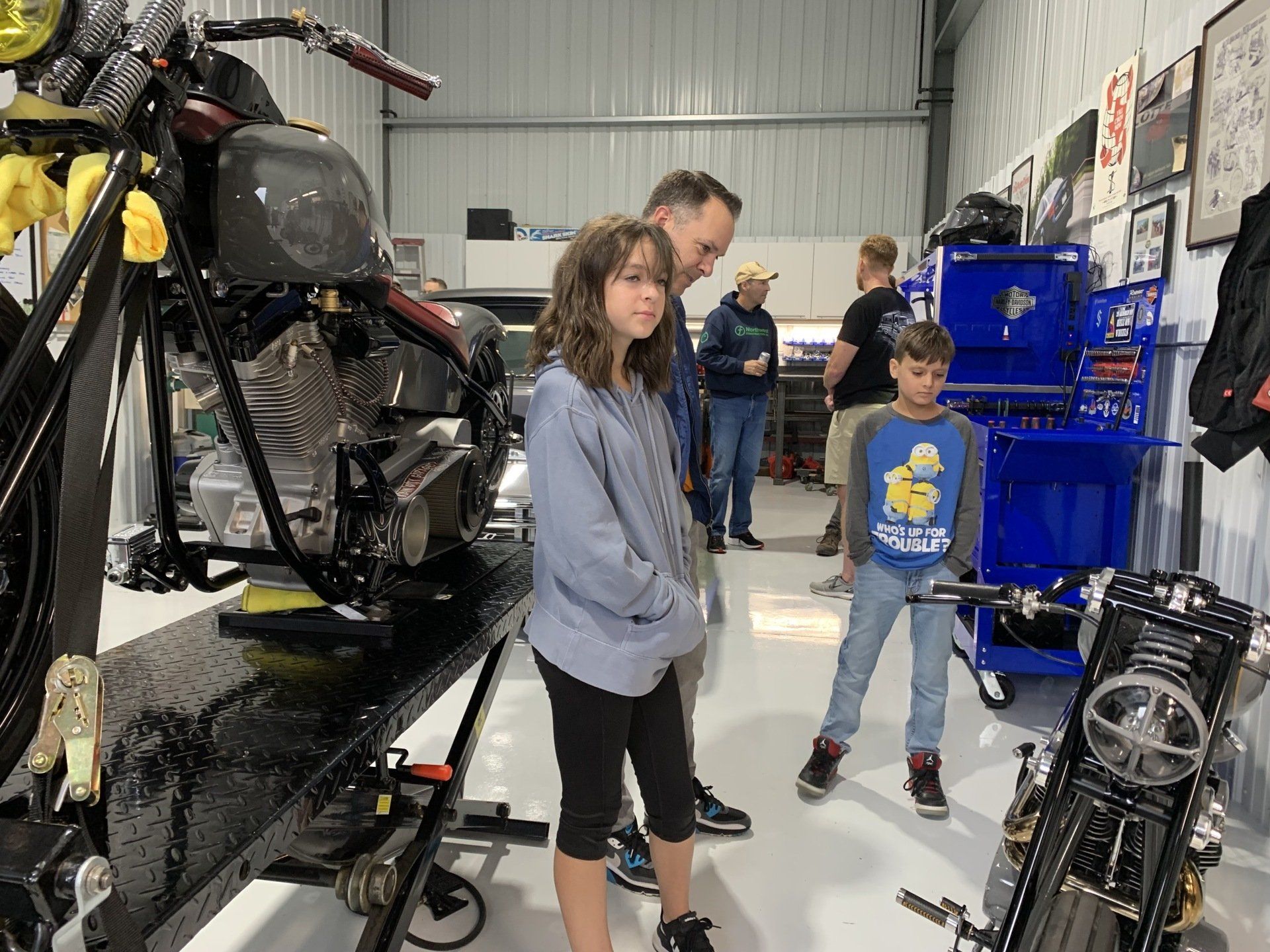 People in a motorcycle workshop looking at partially disassembled bikes and blue tool cabinets.