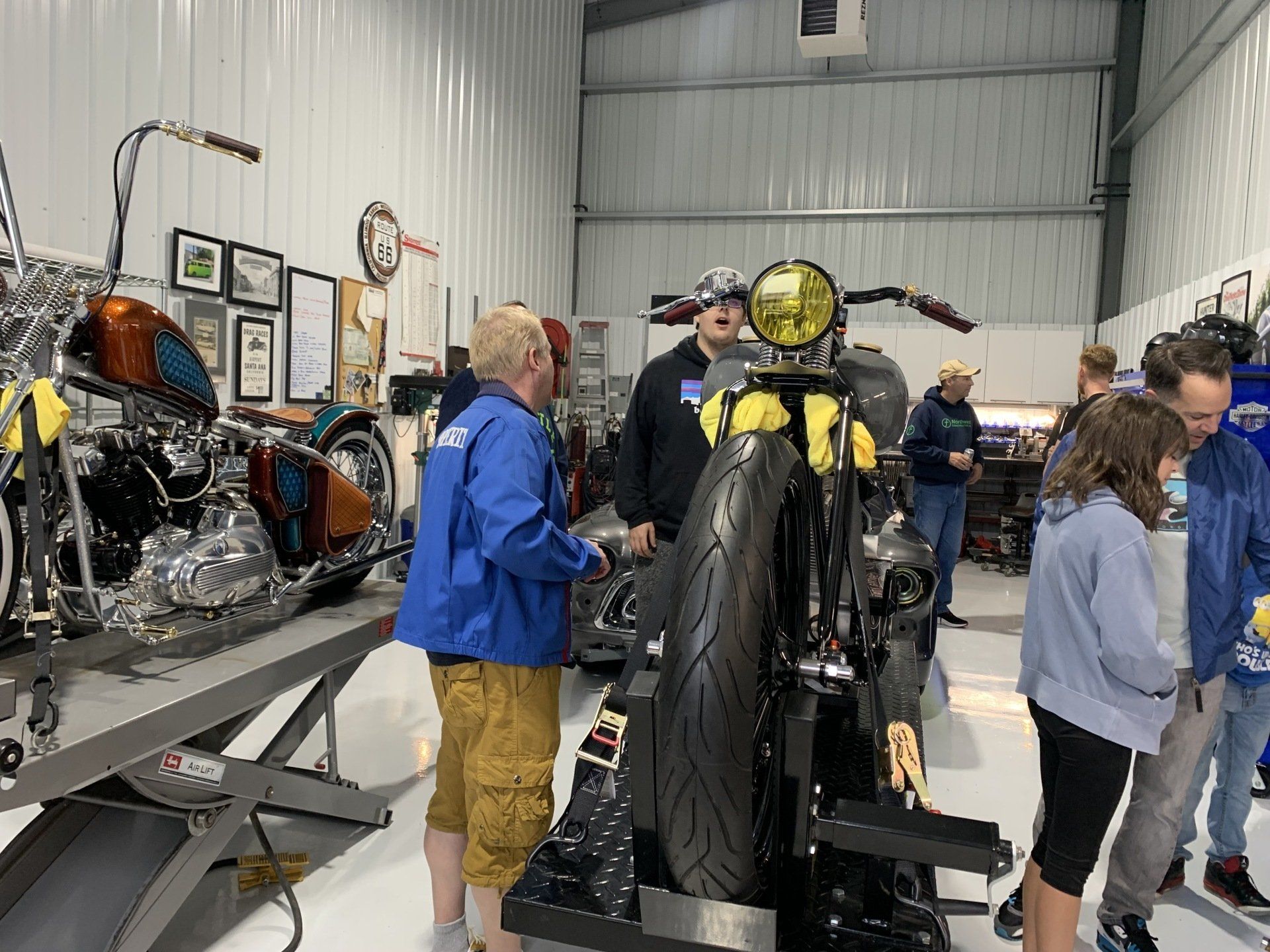 People standing around a large black motorcycle in a garage, with another bike on a lift nearby.