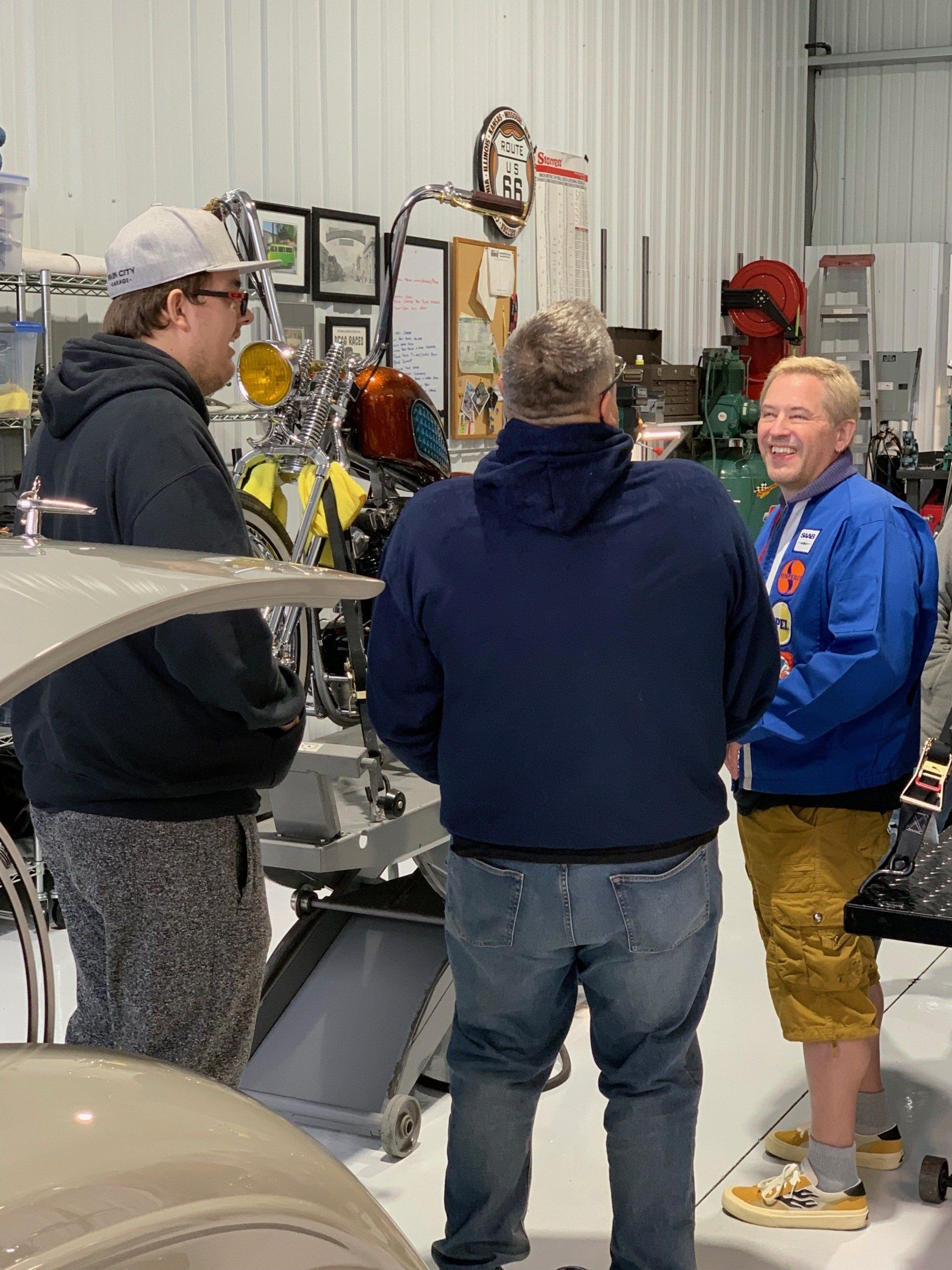 Three people talking in a workshop beside a machine, with tools and shelves in the background.