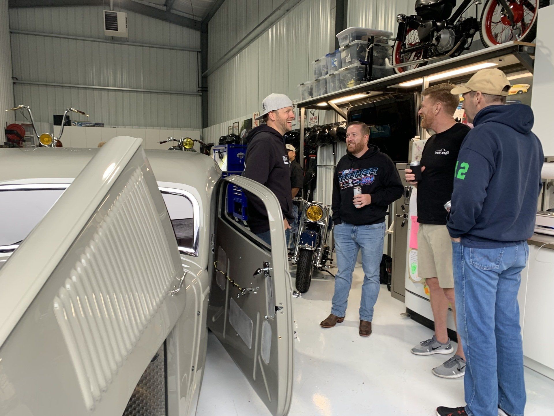 People standing in a garage beside a gray car with its door open, discussing vehicles on a shelf.