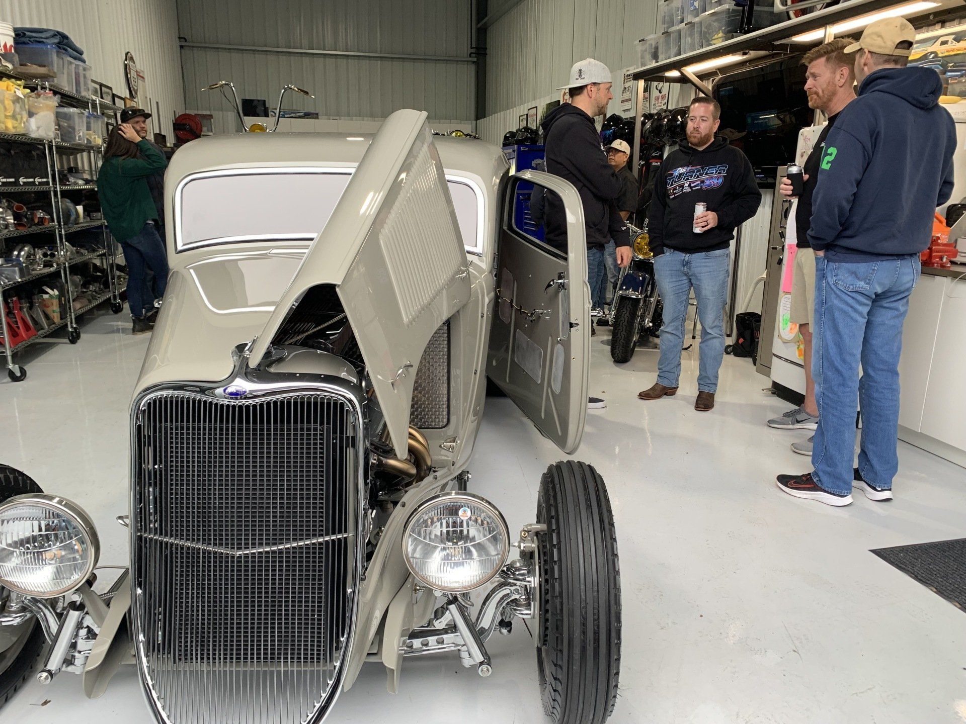Vintage beige hot rod with open hood and doors in a garage, with people standing nearby