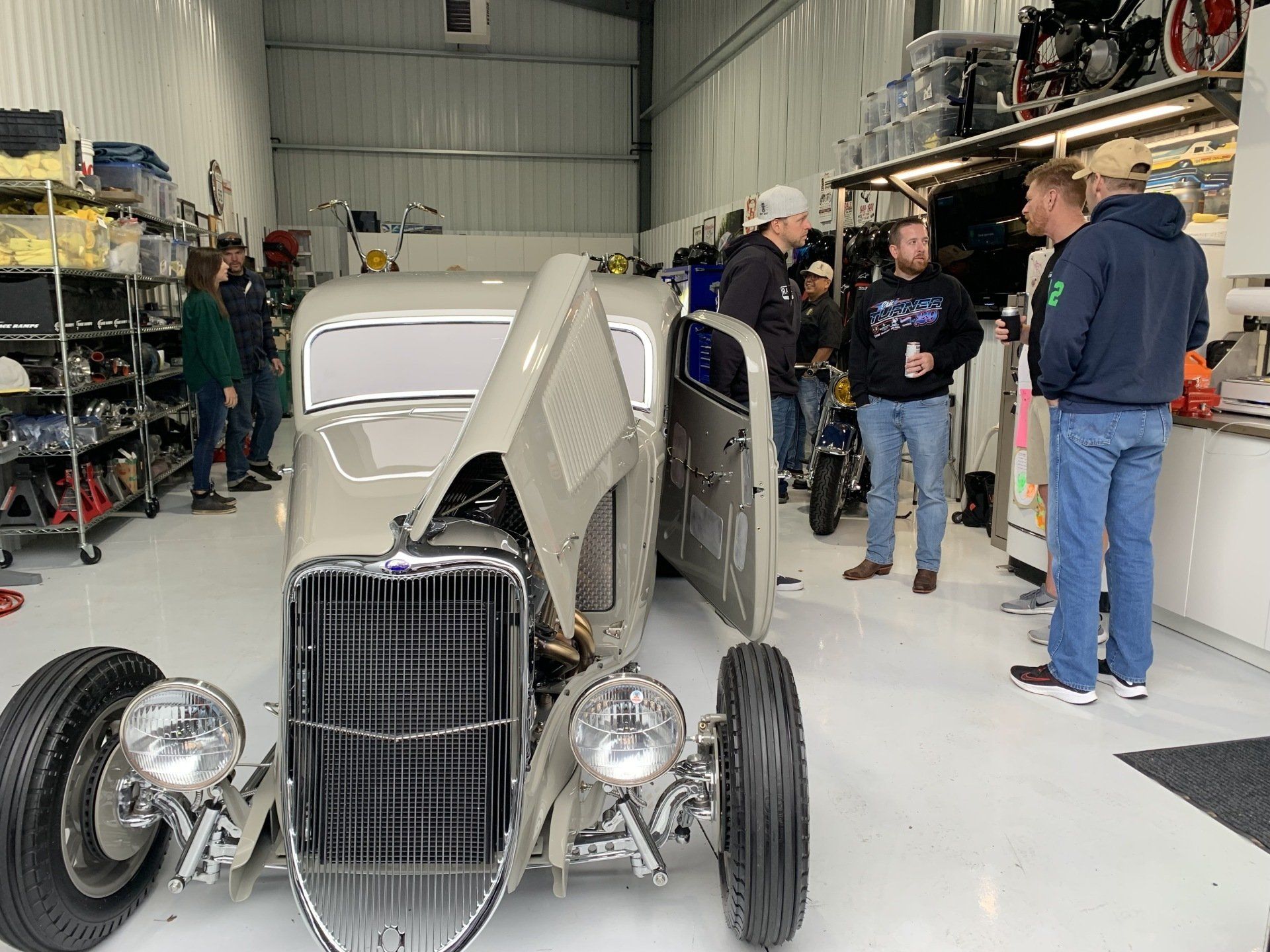 Custom hot rod in a garage with hood open, people standing nearby, and parts on the floor