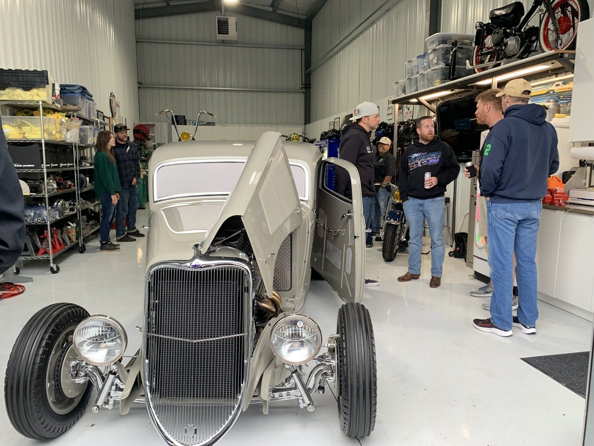 Custom hot rod with open hood in a garage, surrounded by several people