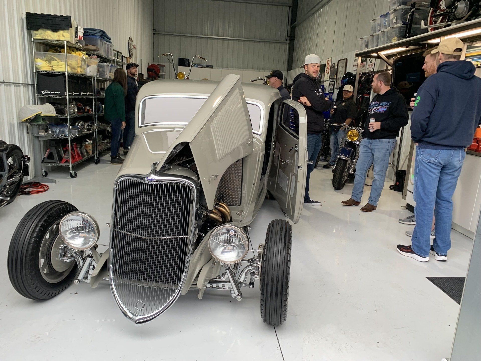 Vintage hot rod with exposed front wheels in a workshop, surrounded by several people.
