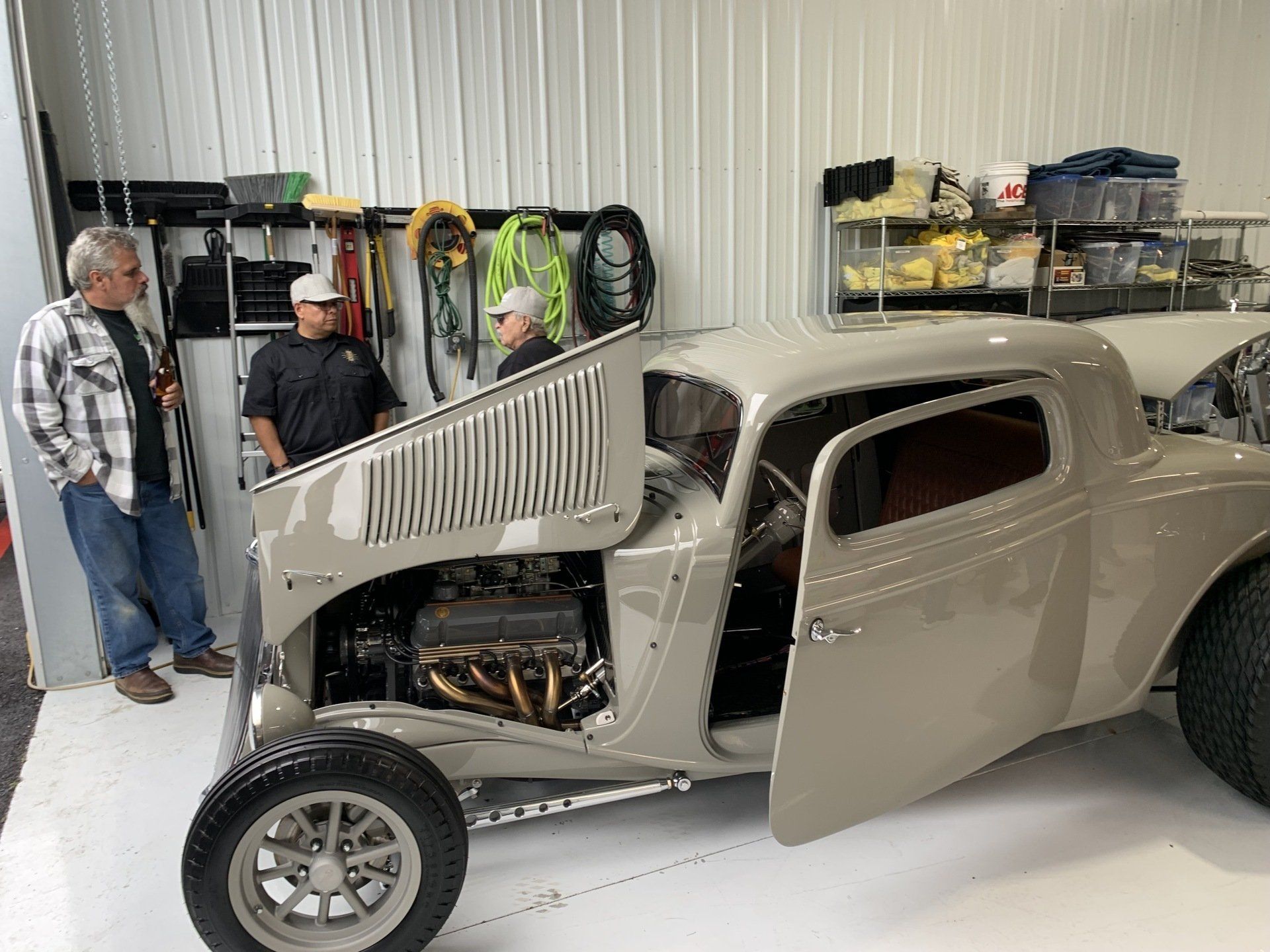Two men inspecting a beige vintage hot rod in a garage with the hood open