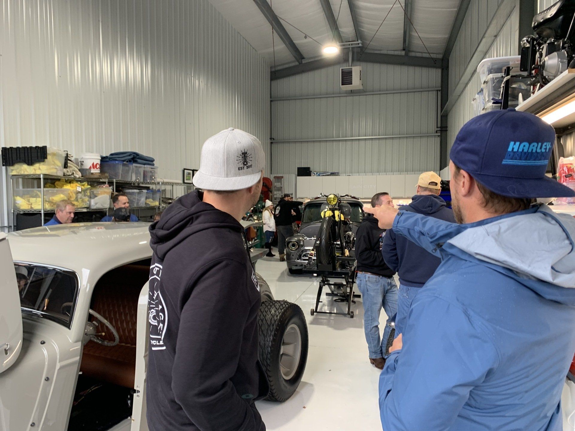 Two people in a workshop point toward a race car inside a metal garage.