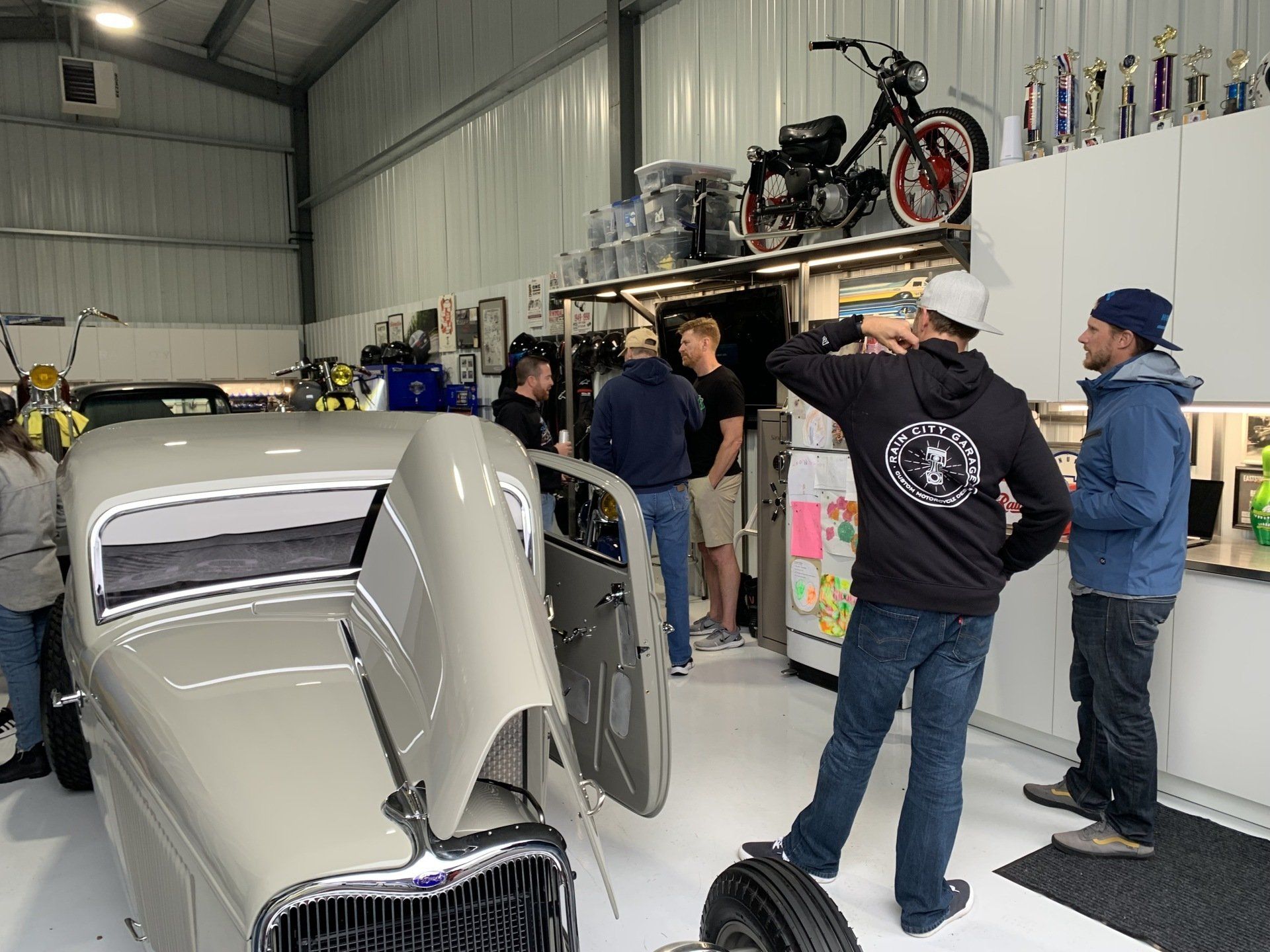 People gathered in a garage beside a silver car, with motorcycles displayed overhead.