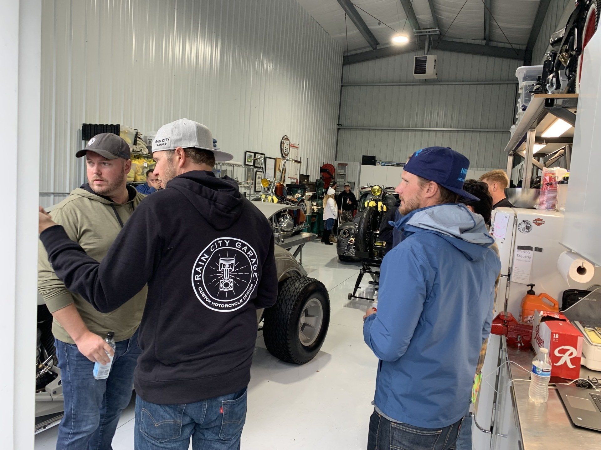 Three people talking in a workshop beside a small off-road vehicle and tools on shelves.
