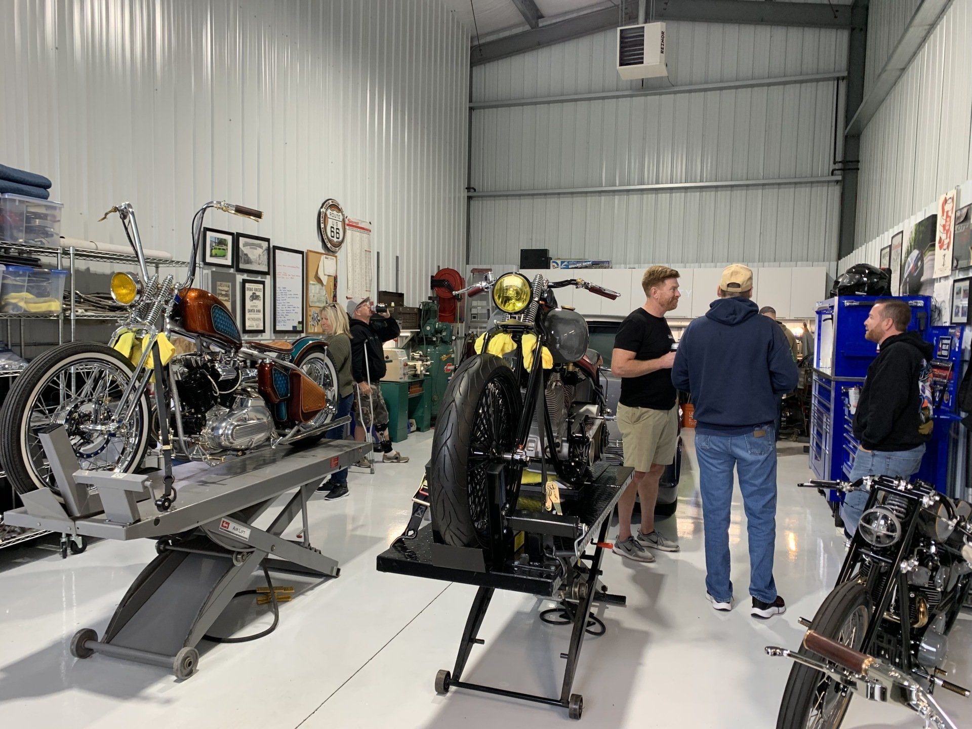 People inspecting custom motorcycles in a bright workshop with white walls and metal stairs