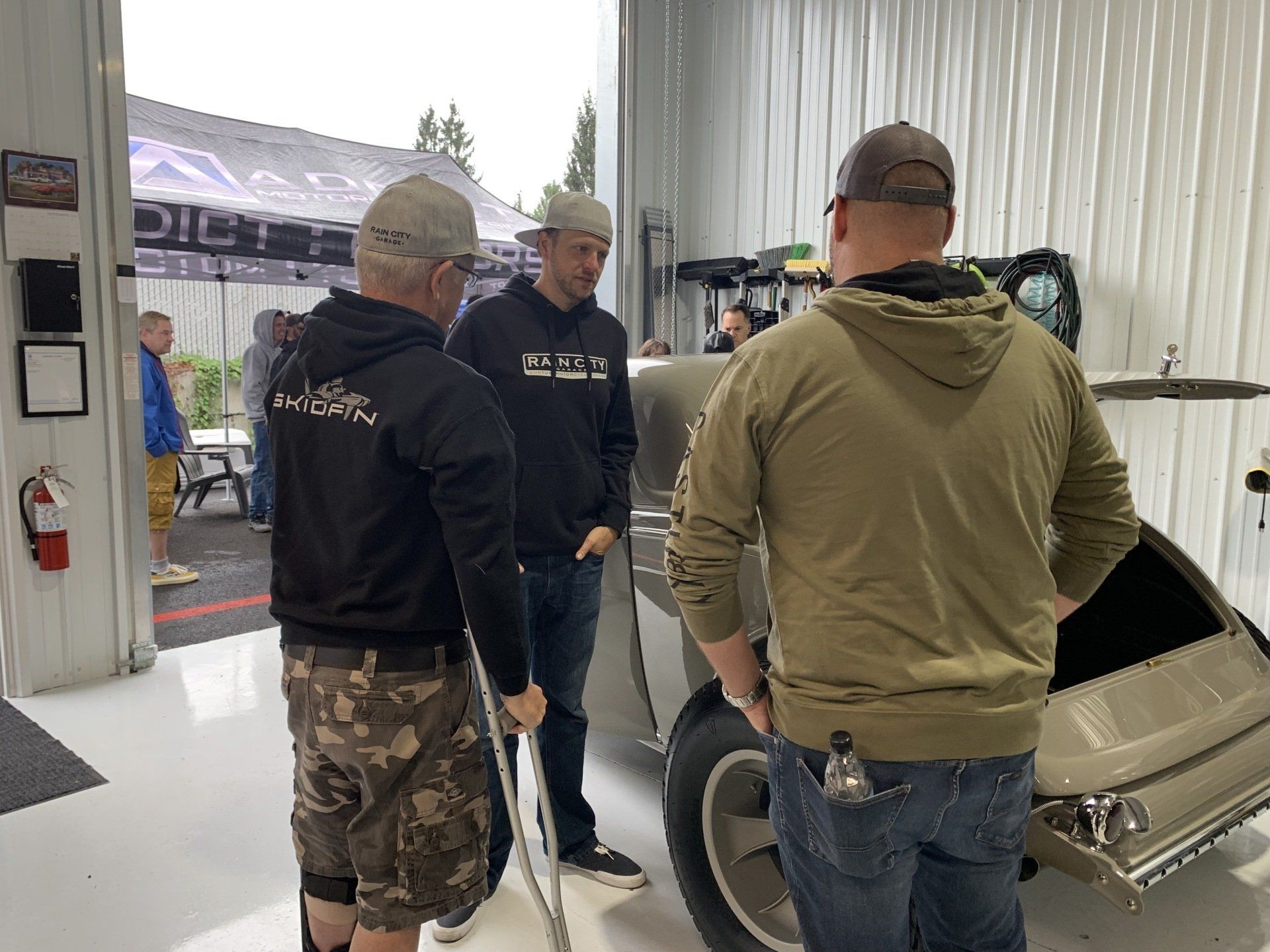 Three men talking beside a small trailer in a workshop-like garage.