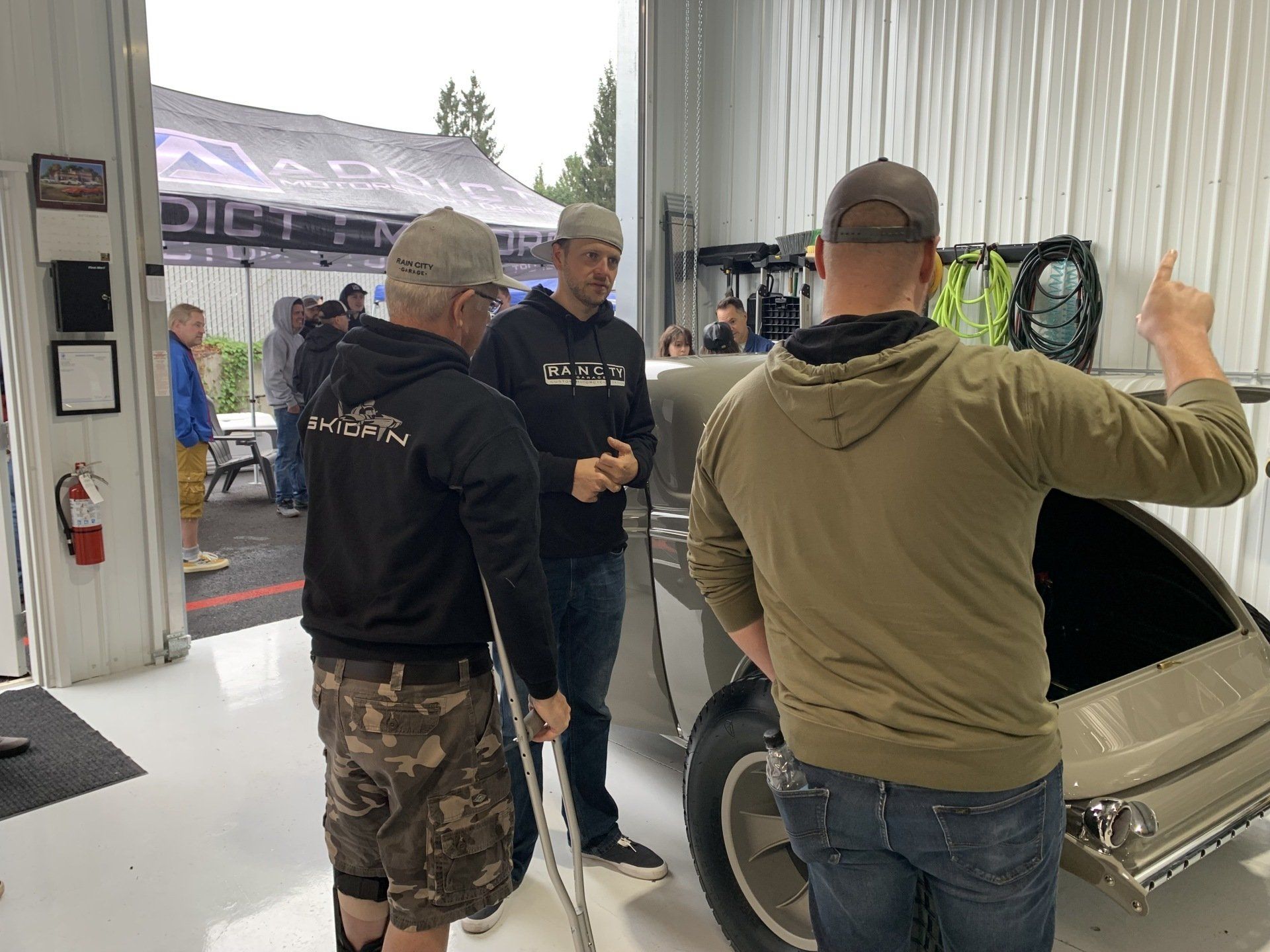 Three men talk in a garage beside a silver car, with one pointing toward the hood.