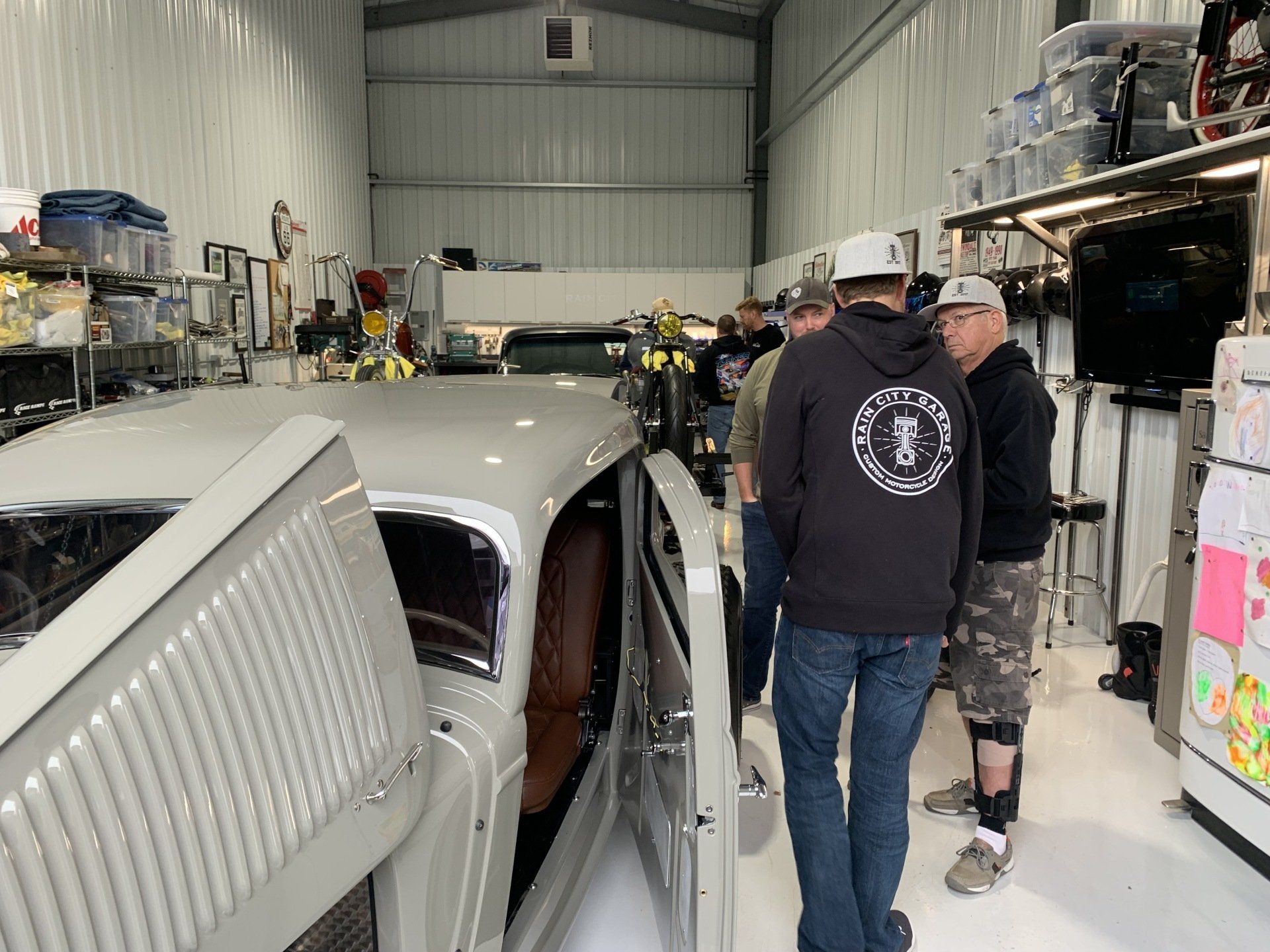 People standing beside a classic car in a garage, with the car hood open and shelves in the background