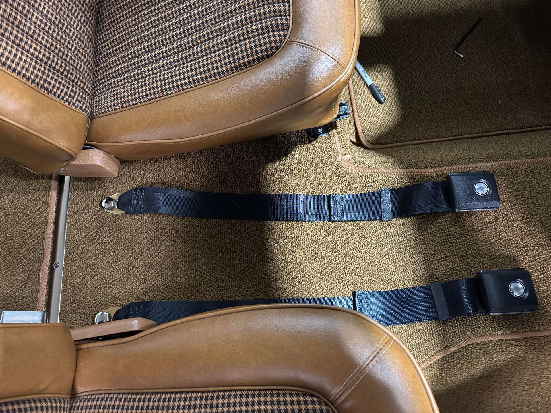 Two brown seats with black seat belts laid across tan carpeted flooring in a vehicle interior