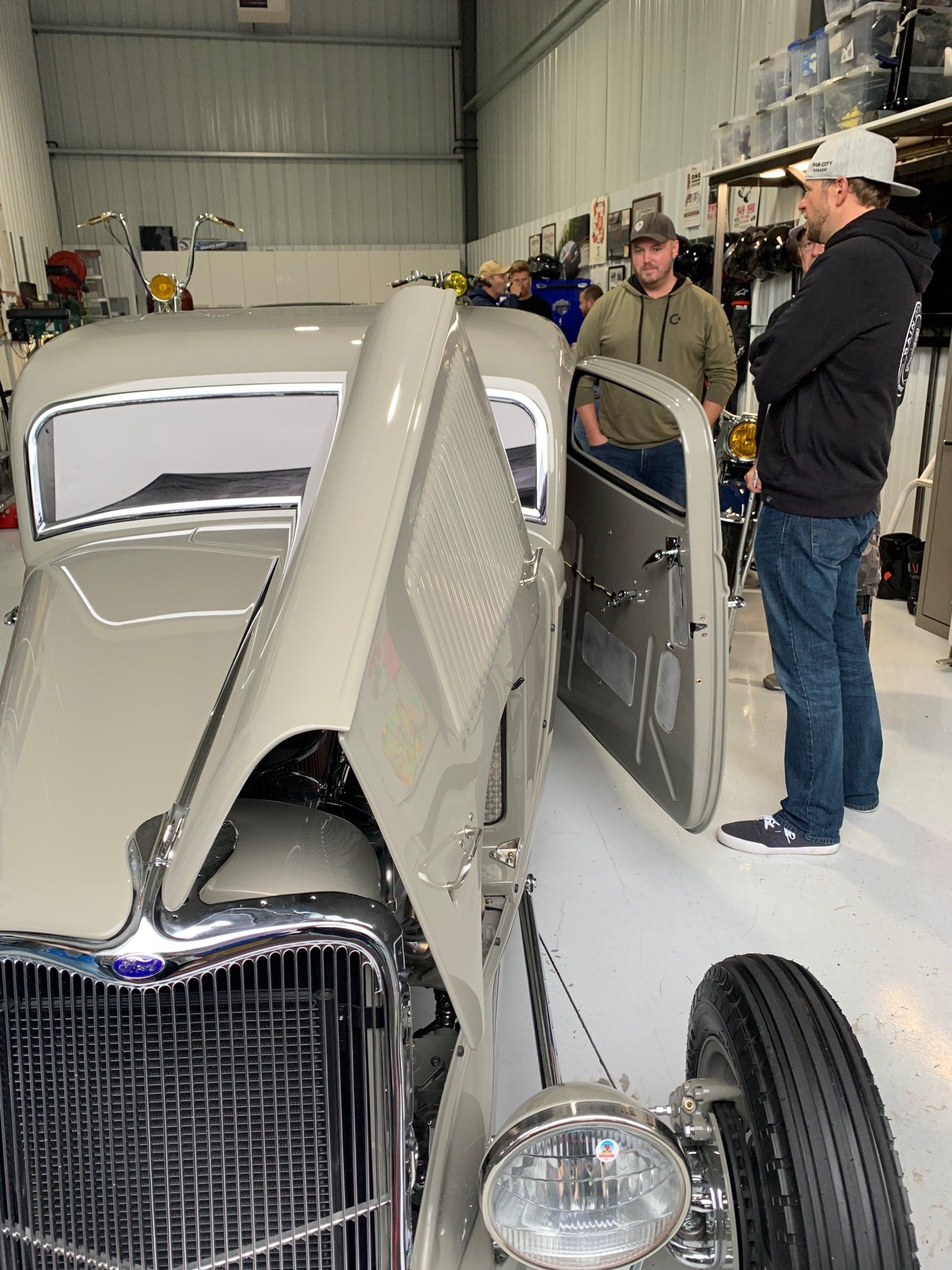 Vintage car in a garage with its hood open while two people stand beside it.