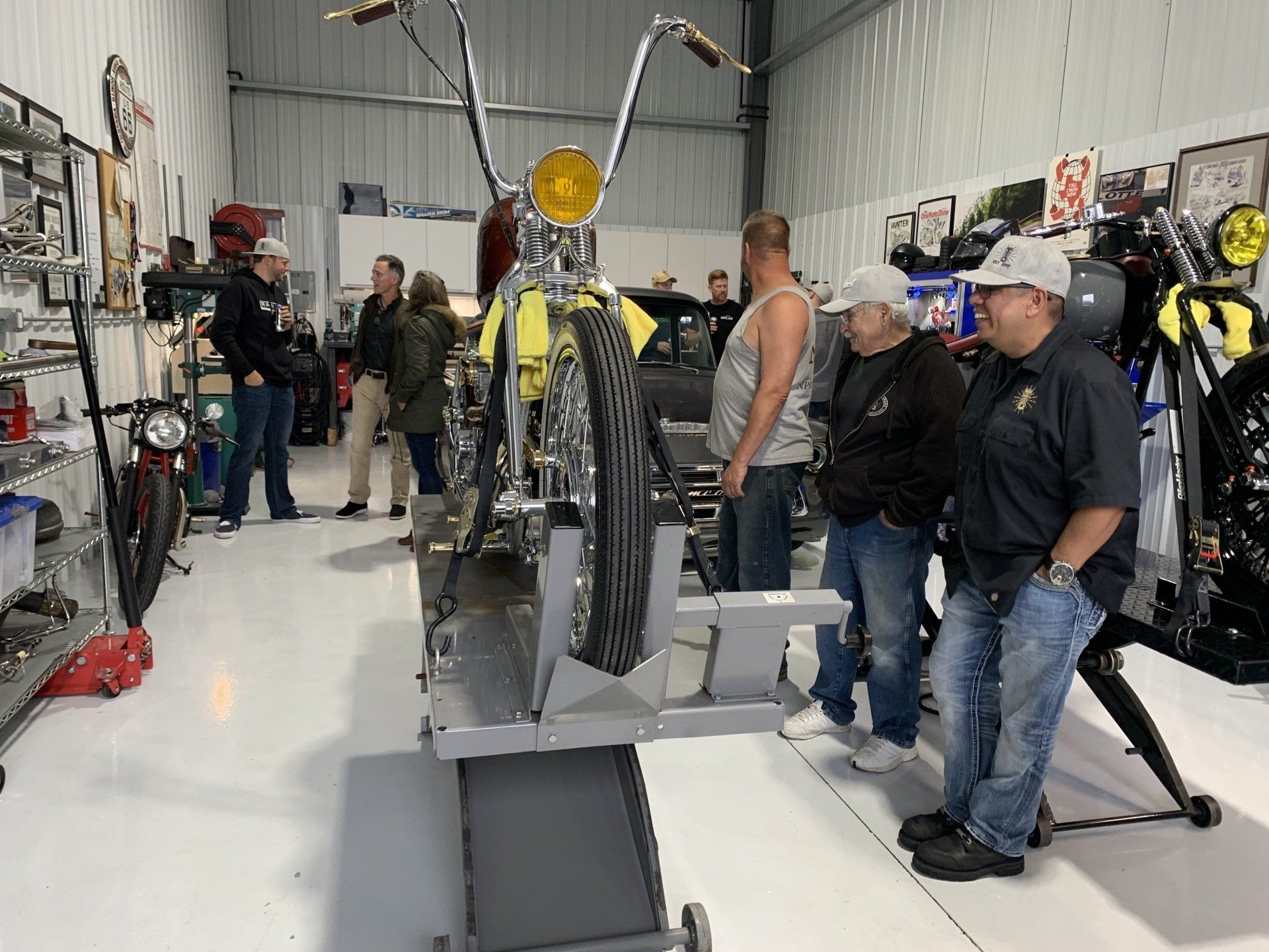 People gathered around a motorcycle in a workshop, with the bike centered on a lift.