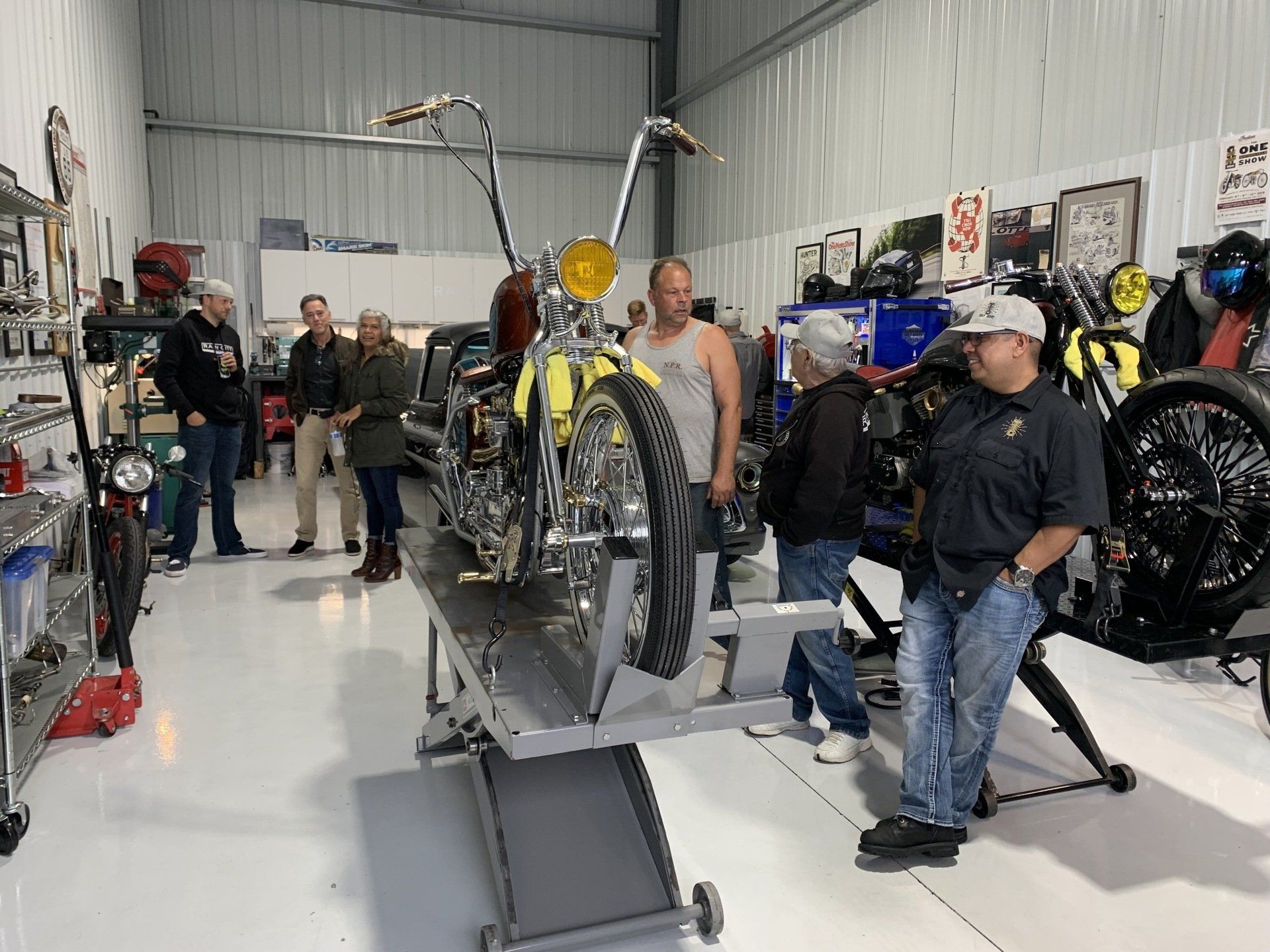 Motorcycles displayed in a workshop; several people stand around a bike on a lift.